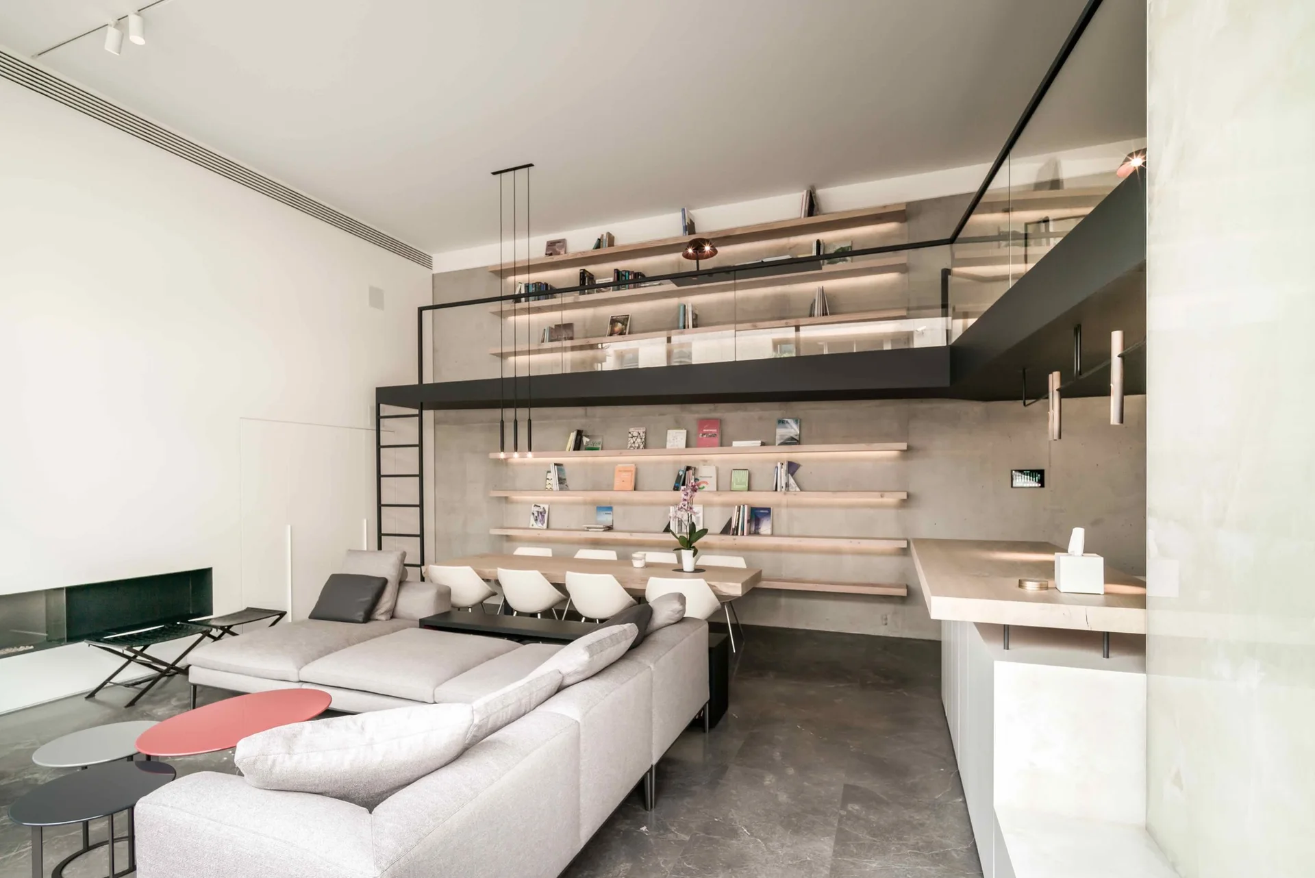 A modern open-concept living space features a gray sectional sofa, a wooden dining table against a concrete wall, and a lofted mezzanine level.