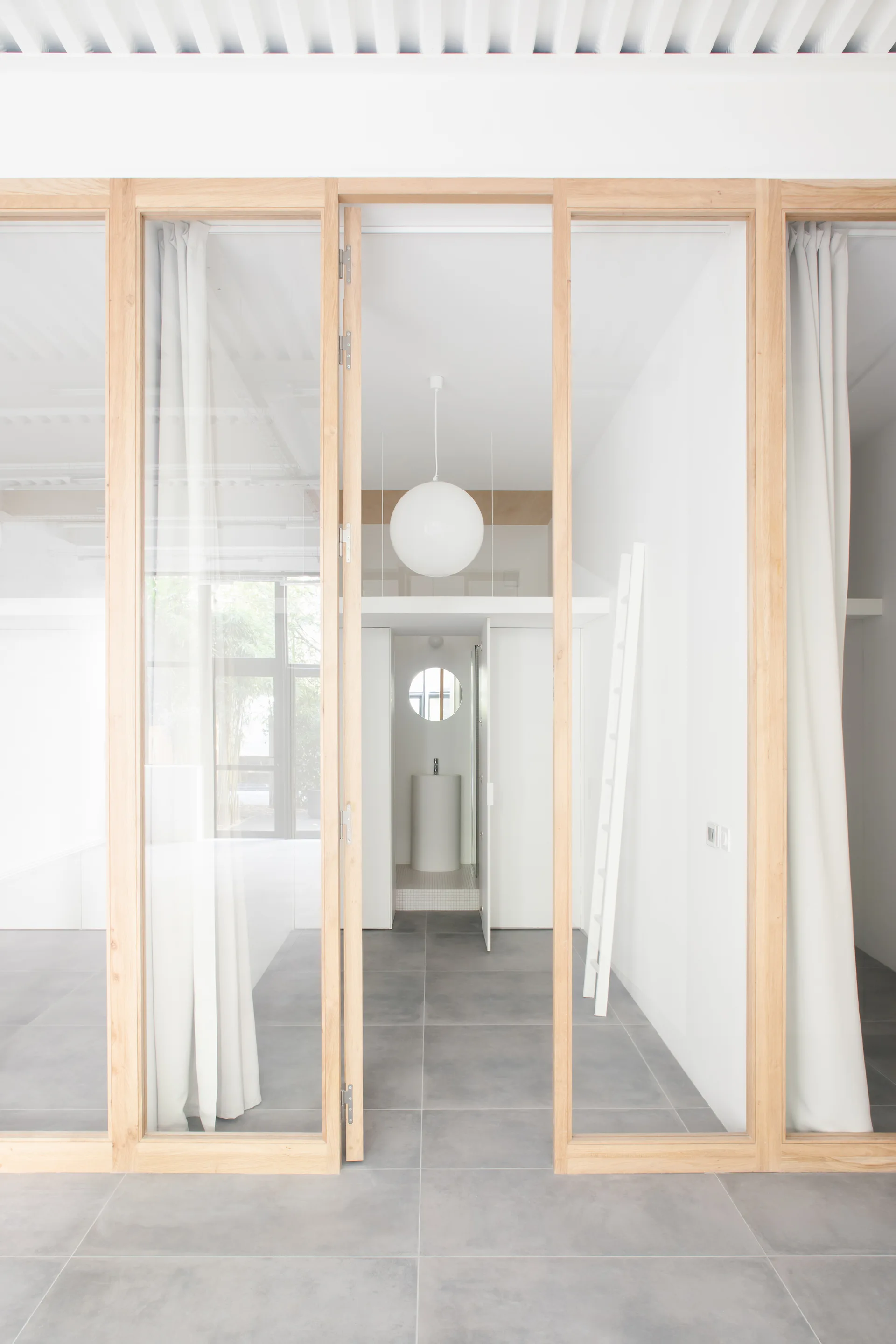 An interior view through a wood-framed glass partition reveals a minimalist white space with grey floor tiles, a sphere pendant, and a loft ladder.