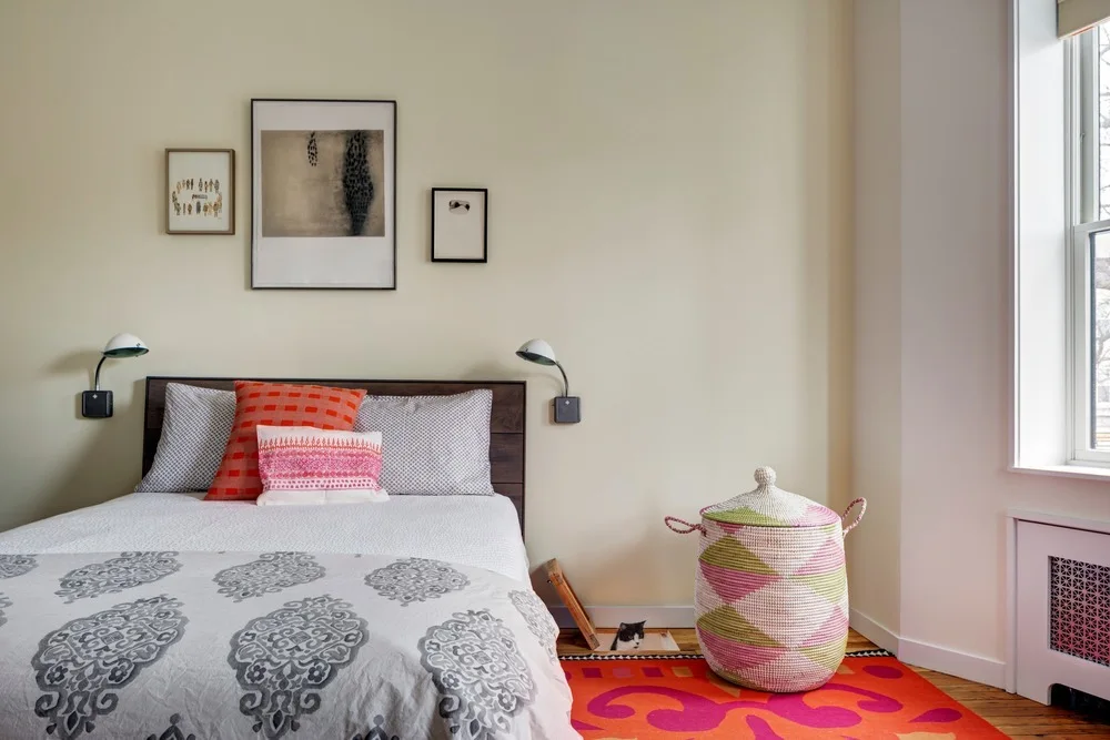 A bedroom features a bed with grey patterned bedding, orange and pink pillows, wall-mounted lamps, and a colorful woven hamper on a vibrant rug.
