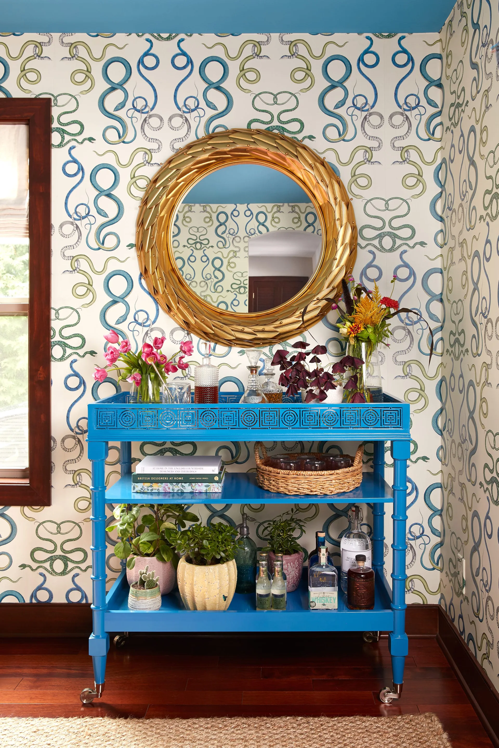 A bright blue three-tiered bar cart styled with plants and decanters stands against green and blue snake-patterned wallpaper under a round gold mirror.