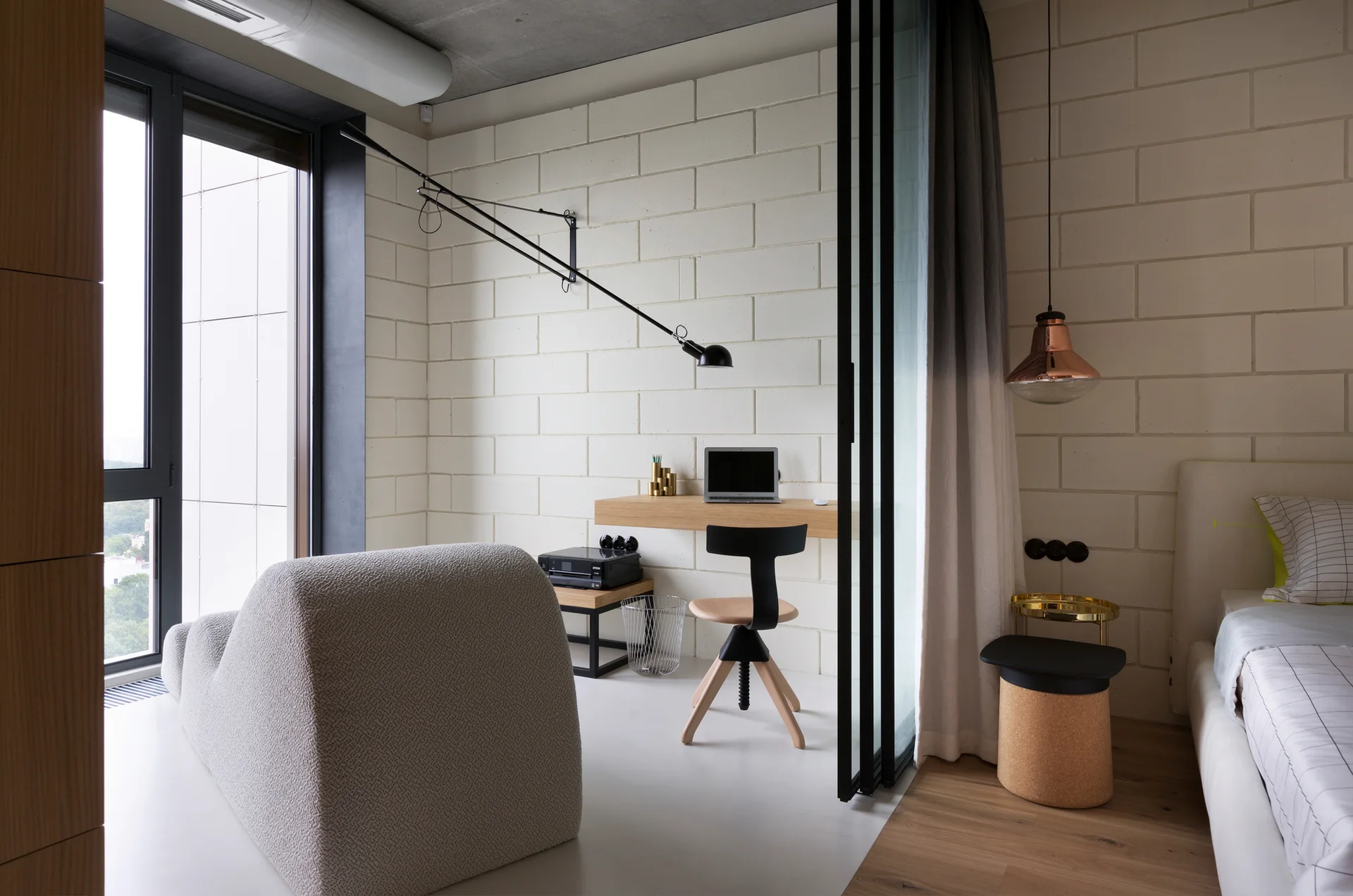 A modern bedroom workspace features a wall-mounted wooden desk, a black swing-arm lamp, and an exposed painted block wall next to a sliding glass partition.