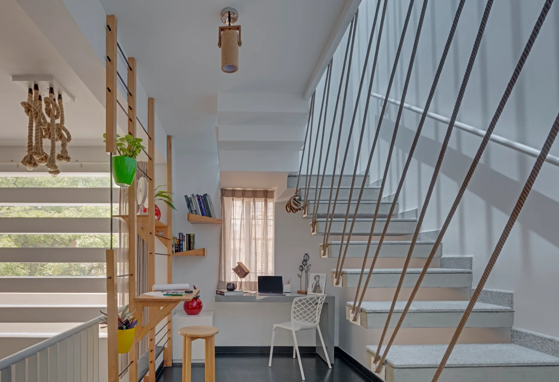 A modern home office built under a concrete staircase featuring a vertical rope balustrade, a grey built-in desk, and light wood shelving.