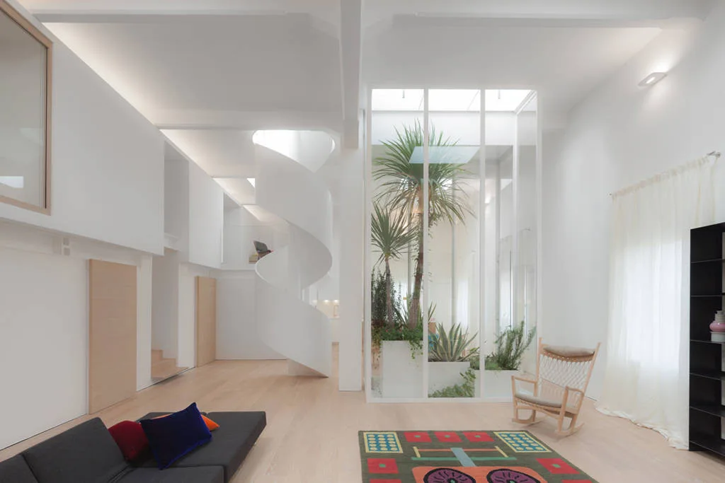 A minimalist living room features a white spiral staircase next to a glass-enclosed plant atrium beneath a skylight.