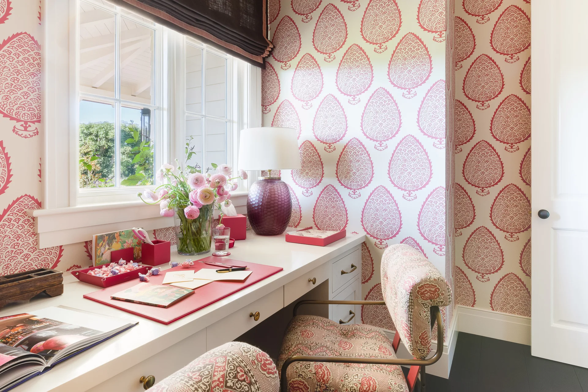 A white built-in writing desk sits in a home office alcove covered in pink and white botanical patterned wallpaper beneath a window.