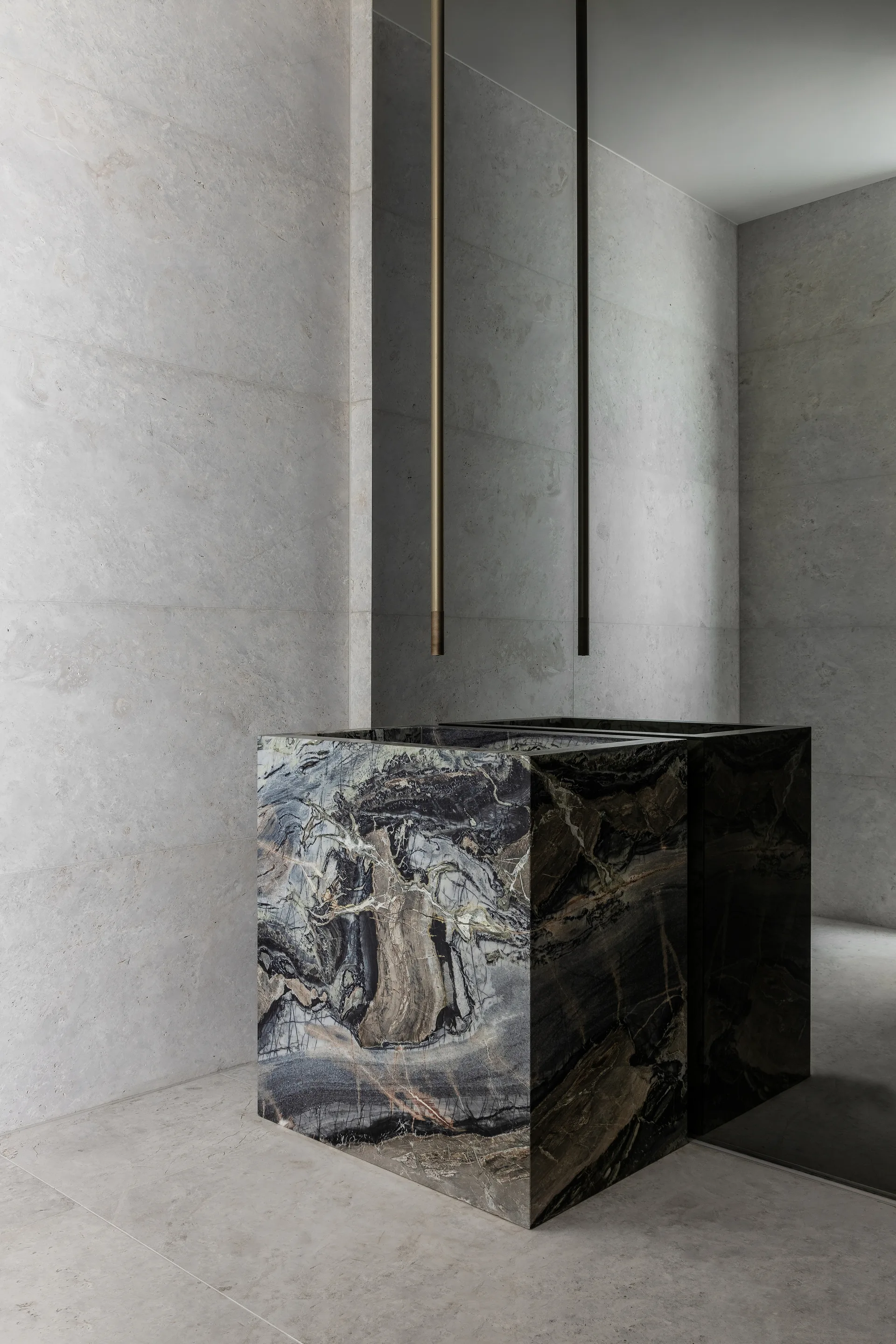 A rectangular dark veined marble pedestal sink stands against a light gray tiled wall beneath two slim vertical ceiling-mounted faucets.