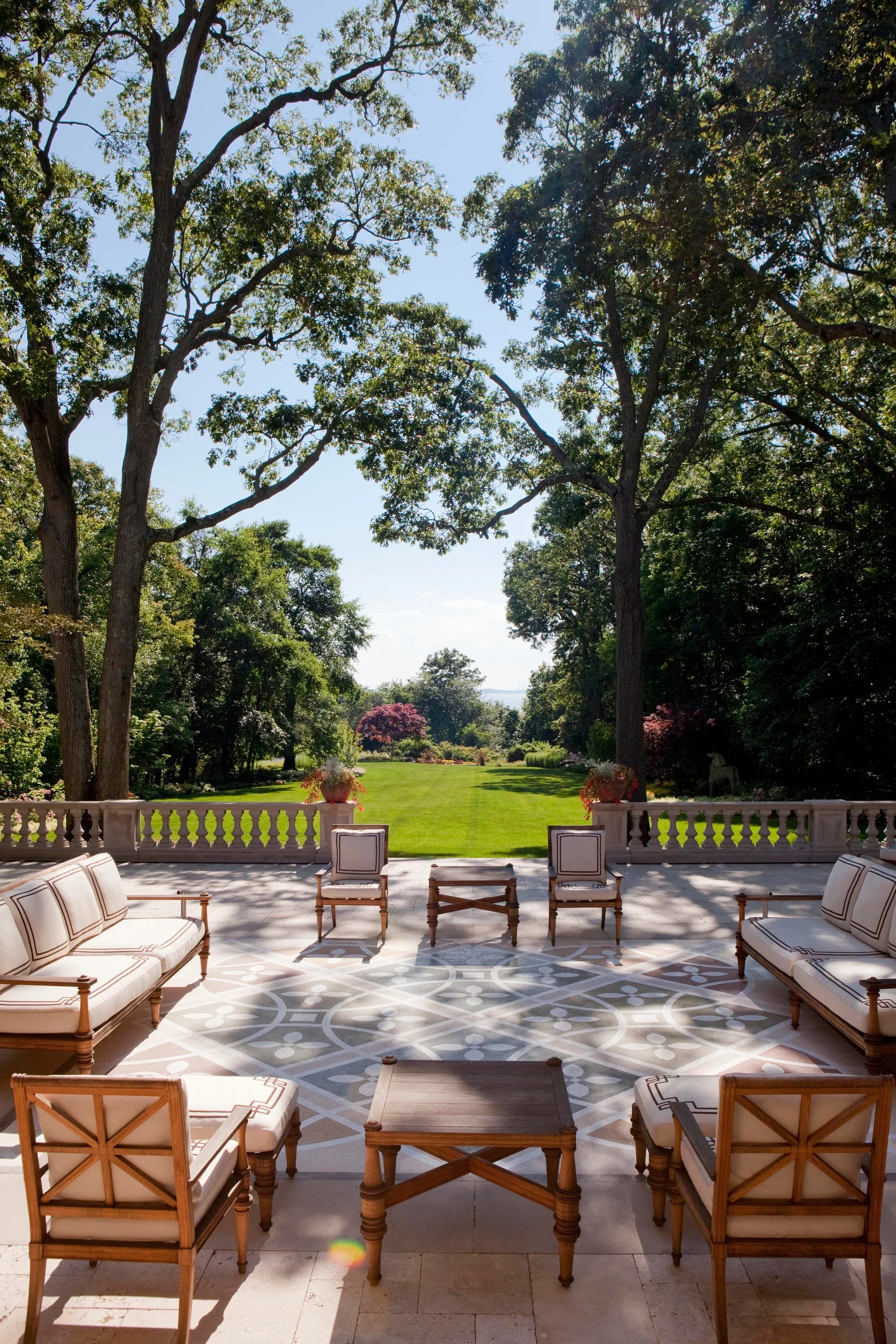 A formal stone patio with symmetrical wooden outdoor furniture and an intricate geometric floor inlay overlooking a vast green lawn bordered by tall trees.