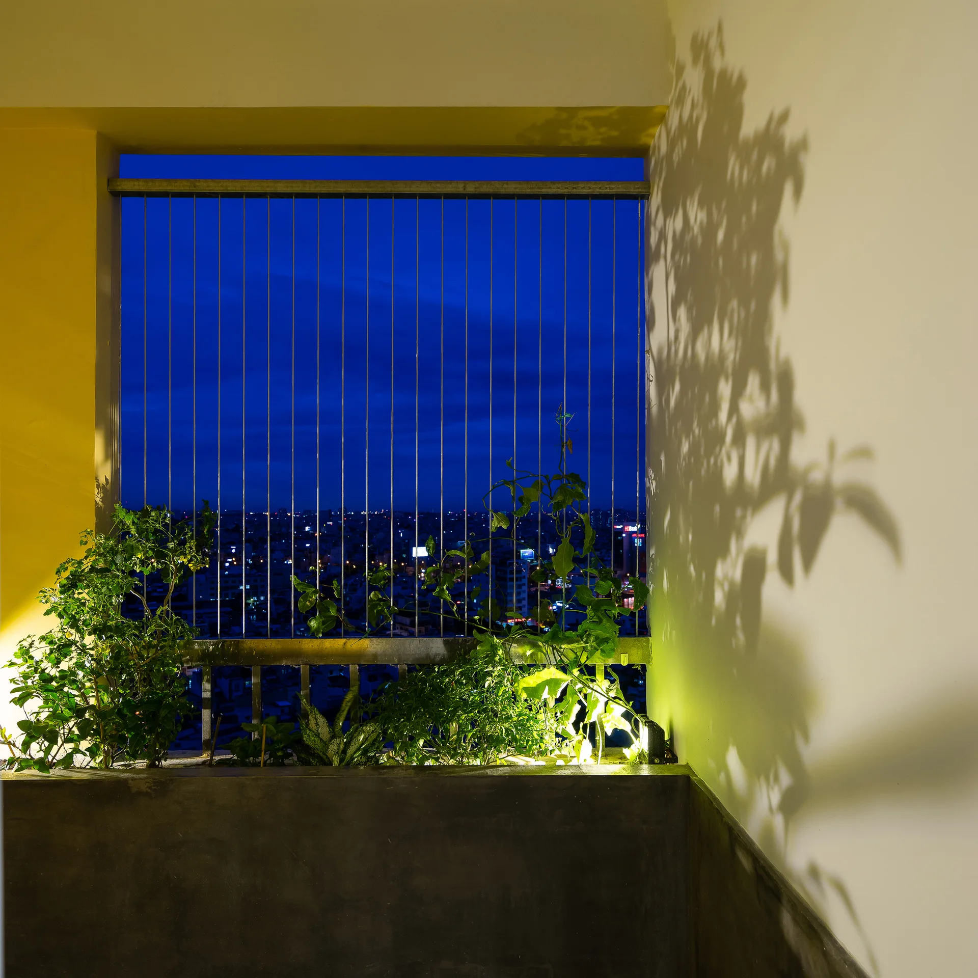 A dark gray concrete planter with illuminated green plants sits on a balcony with yellow walls and a metal railing overlooking a night city skyline.