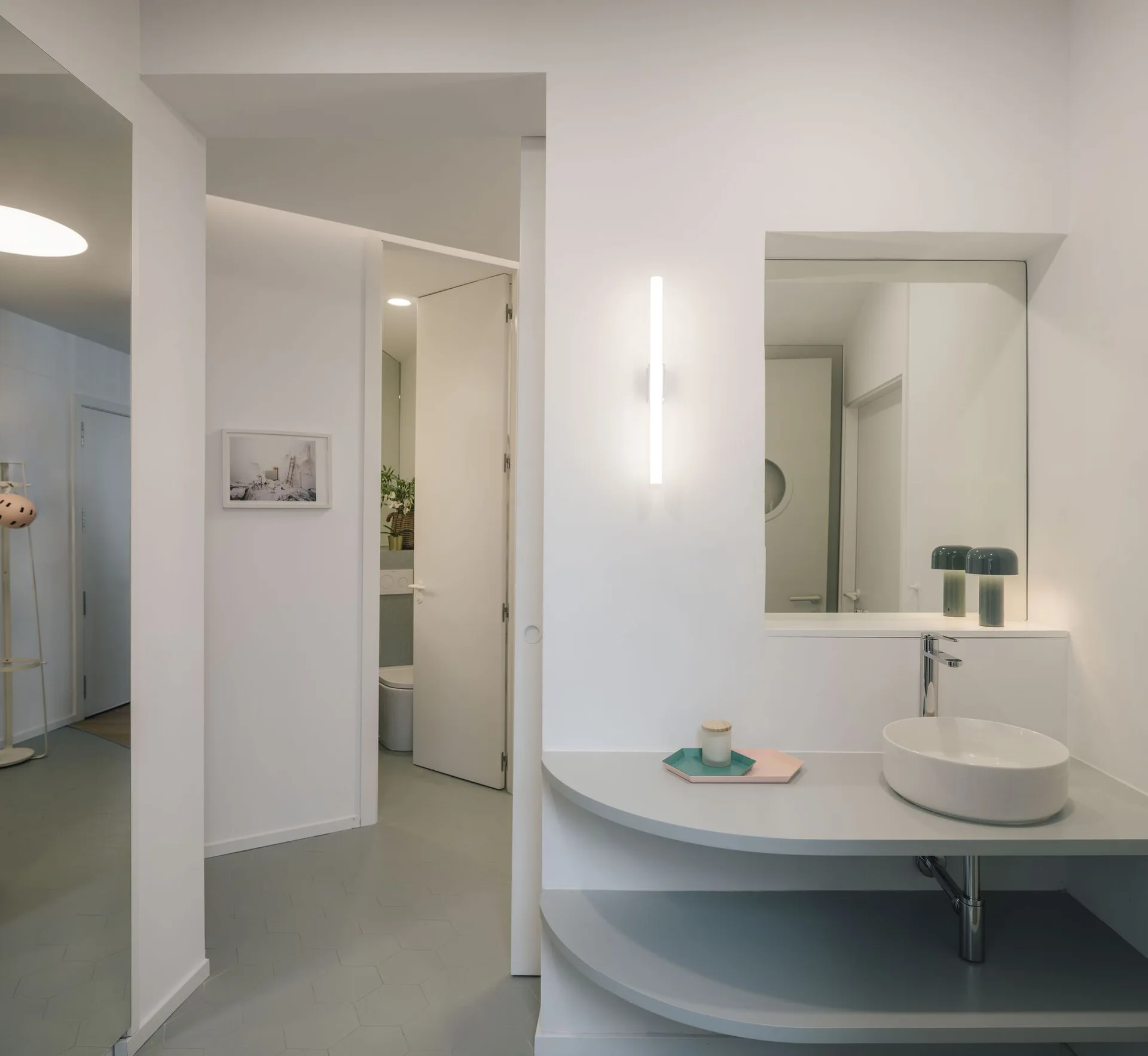 A minimalist white bathroom featuring a curved gray vanity shelf, a vessel sink, a linear wall sconce, and hexagonal gray floor tiles.