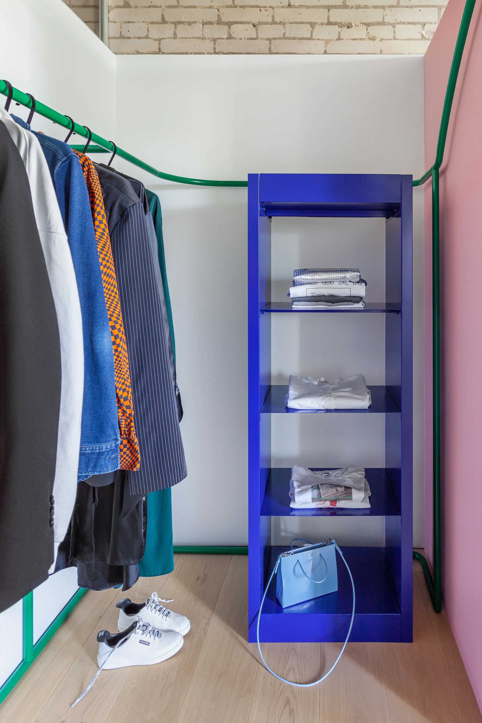 A modern closet features a vibrant blue metal shelving unit and a curved green tubular hanging rod against white and pink walls with wood flooring.