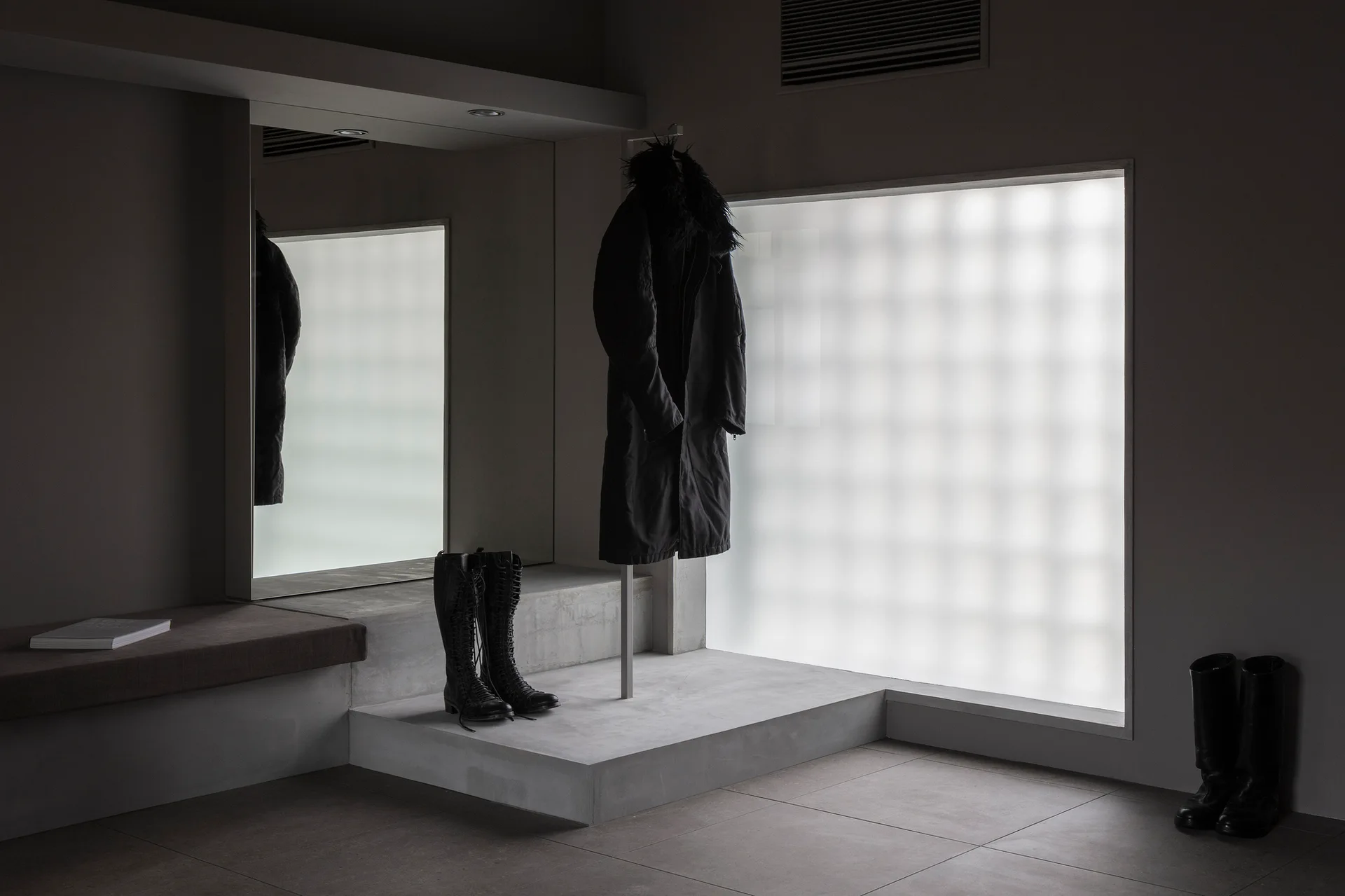 A minimalist gray entryway featuring a raised concrete platform, a large wall mirror, and a frosted glass block window diffusing soft light.