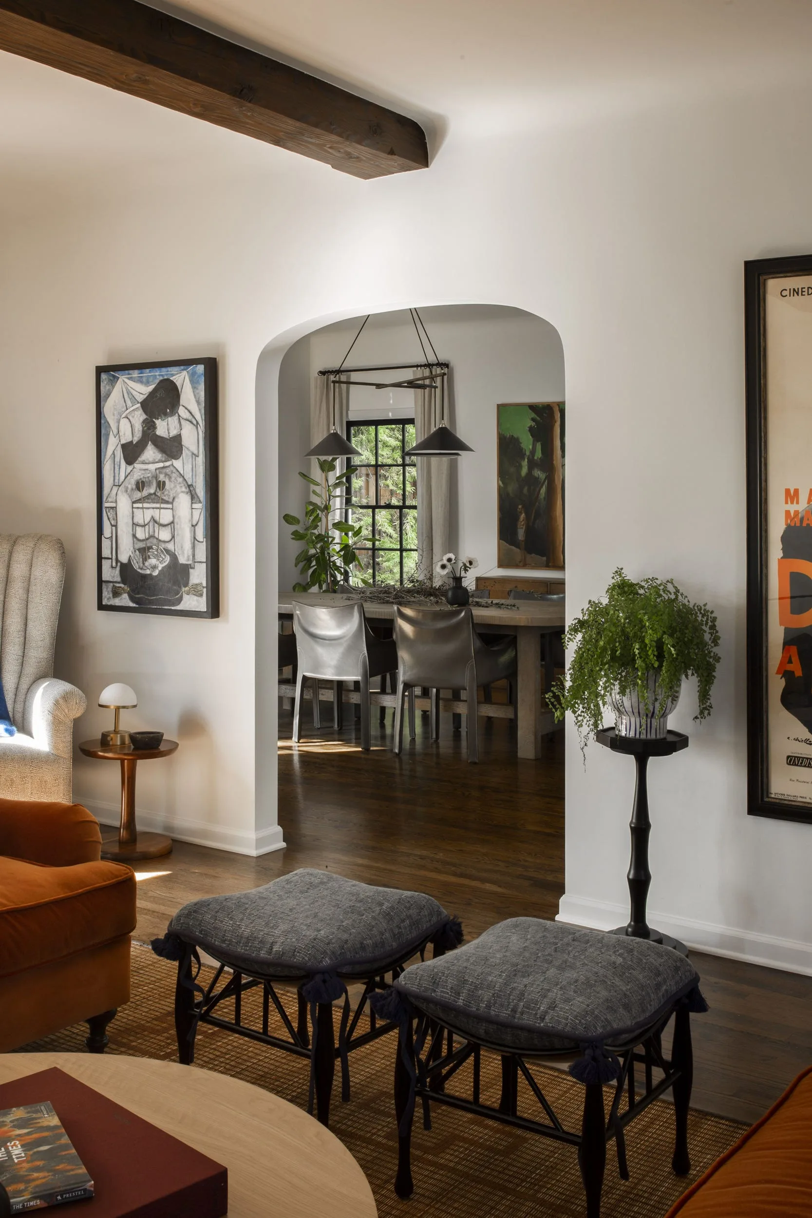 An arched opening links a white living room featuring gray tasseled ottomans to a dining room with a wooden table under dark pendant lights.