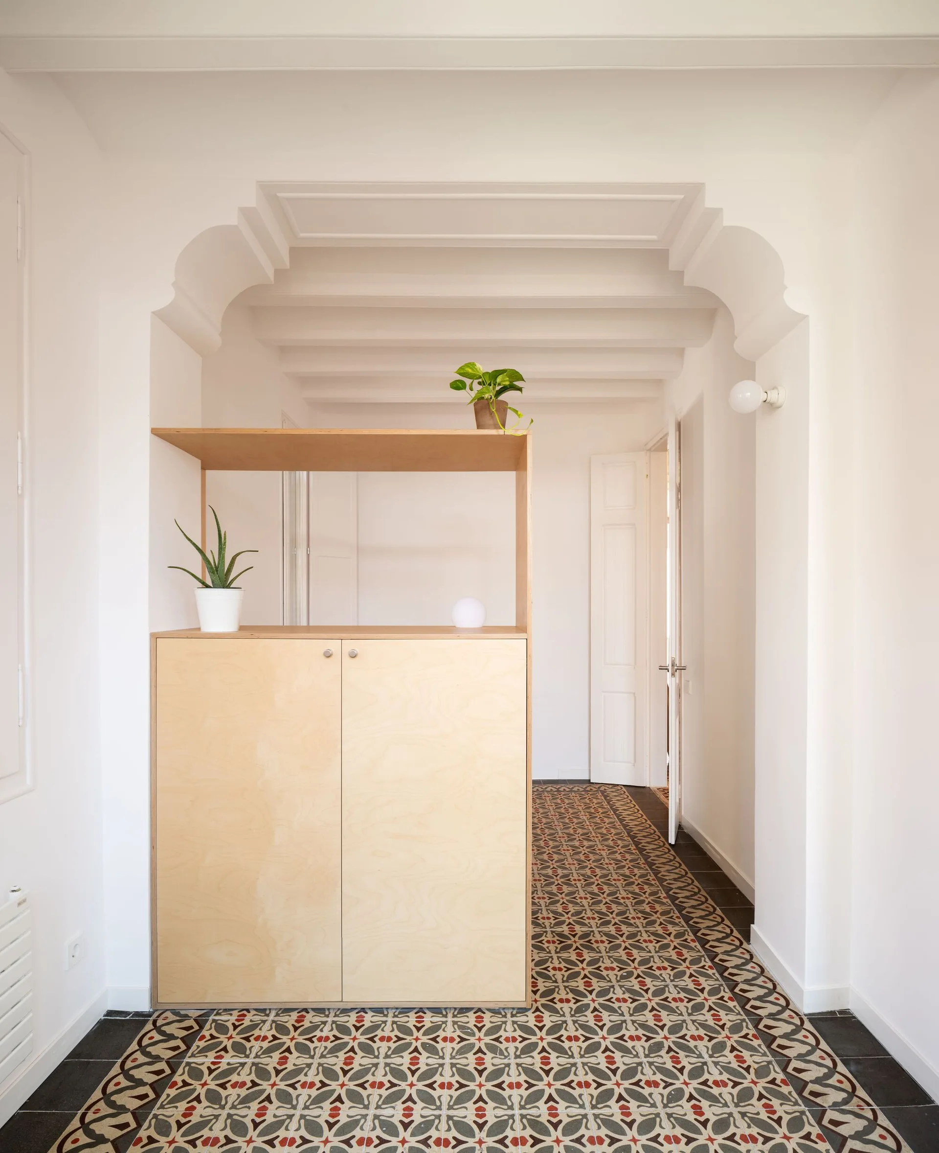 A light wood cabinet divider stands in a white hallway with decorative arched doorways, exposed ceiling beams, and patterned geometric floor tiles.