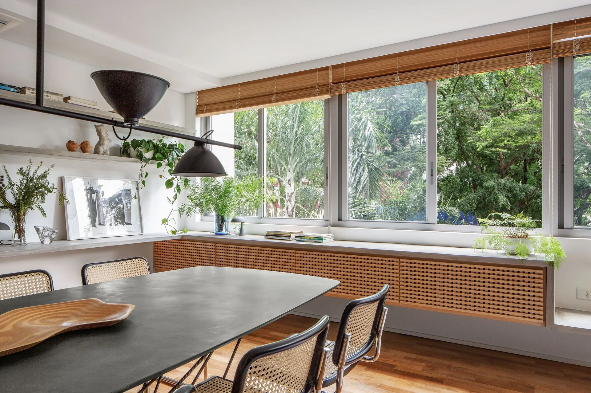 A modern dining room features a dark rectangular table with cane-back chairs, a long wood-grid cabinet under a large window, and floating shelves.