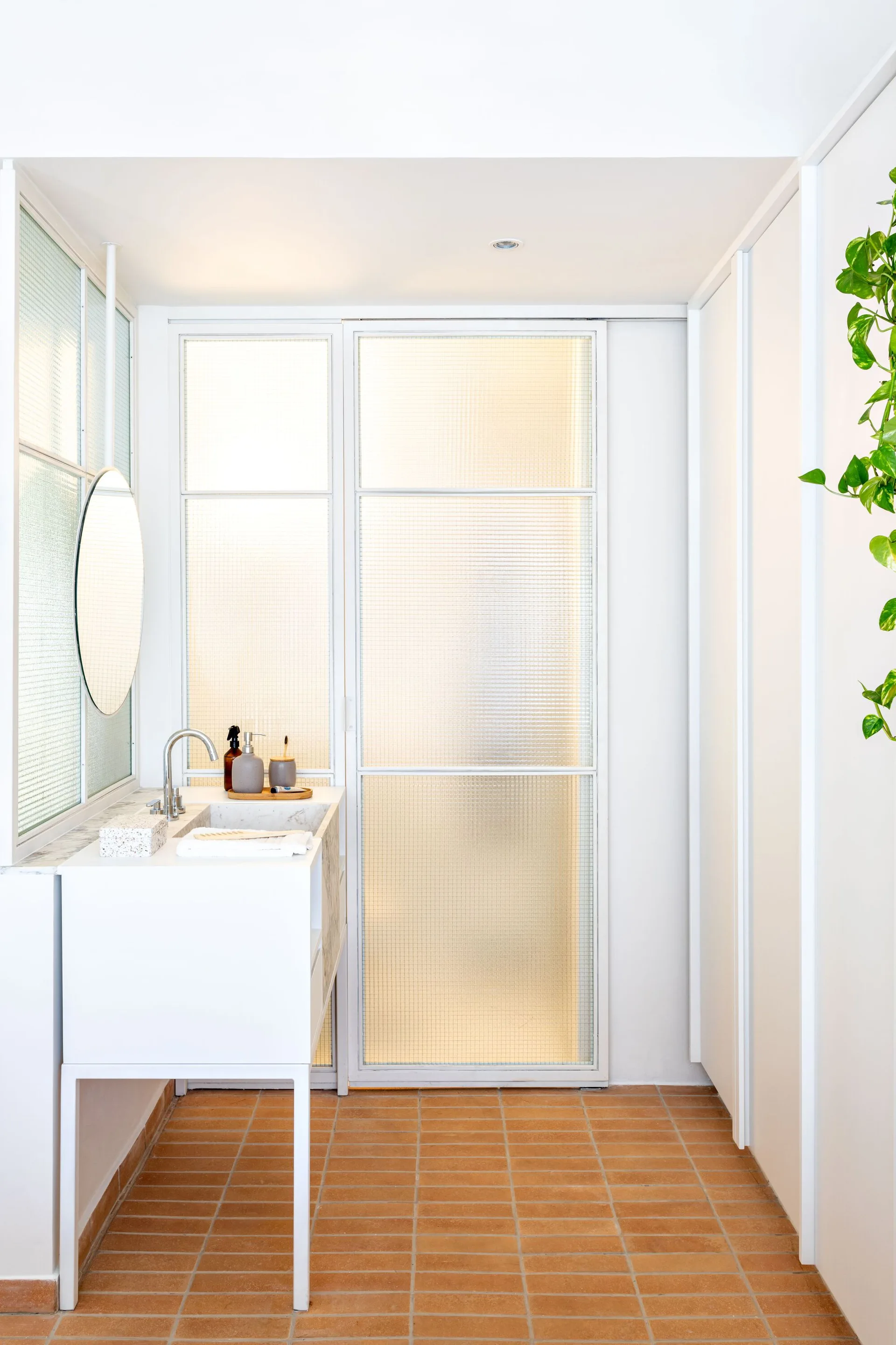 A modern white bathroom vanity with a round mirror is paired with textured glass sliding partition doors and square terracotta floor tiles.