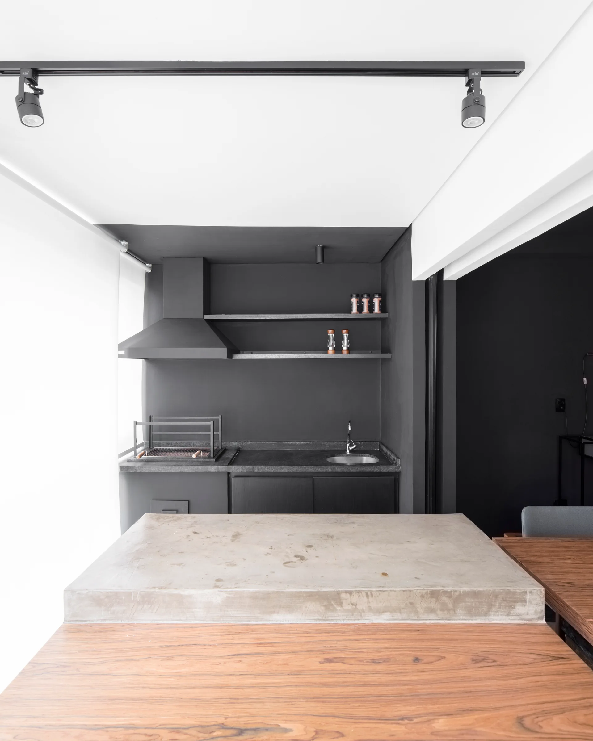 A modern kitchen featuring a concrete-topped island and wooden table in the foreground, with a matte black prep wall, integrated grill, and open shelving.
