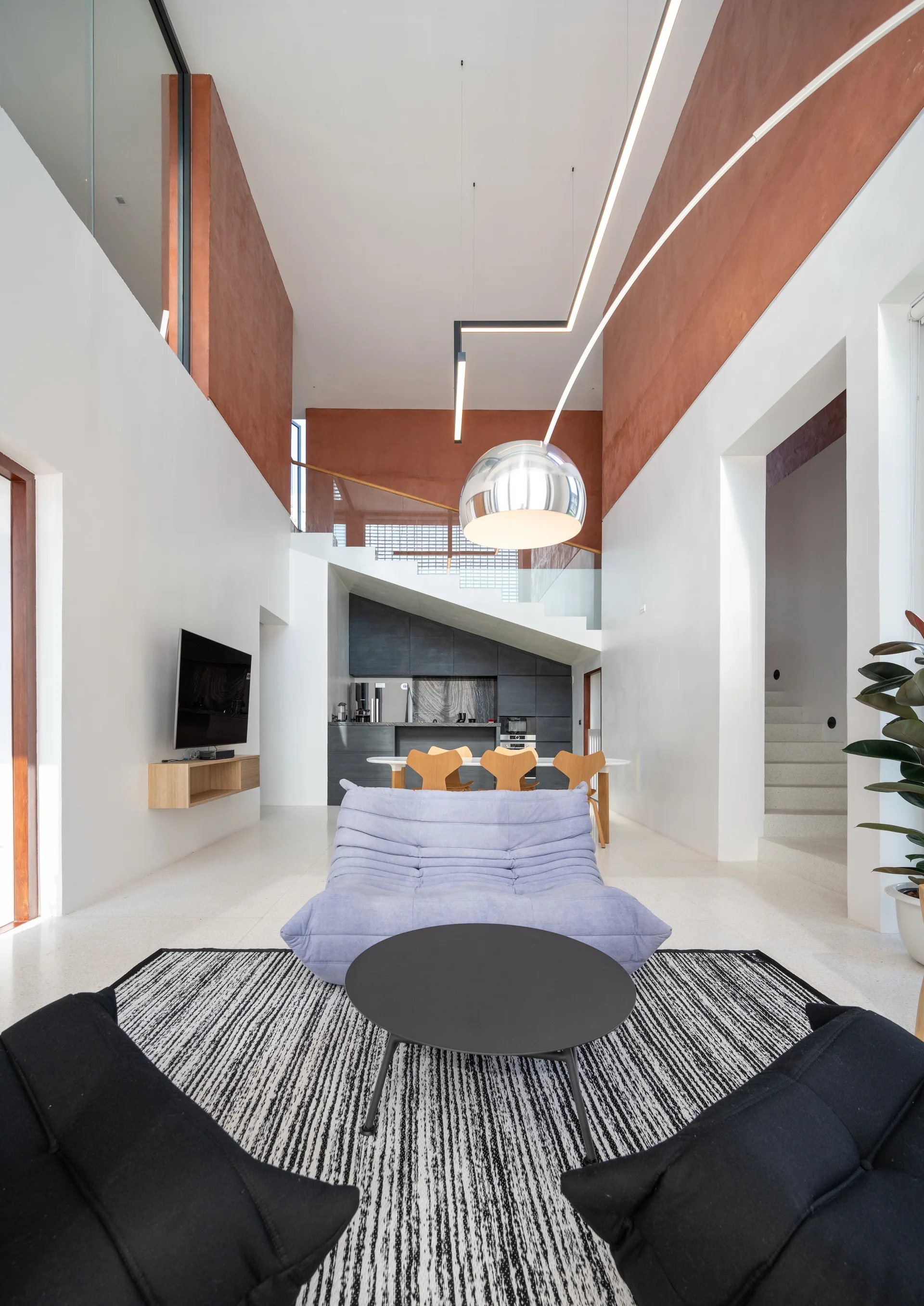 A double-height living room features a purple tufted sofa, a striped area rug, white walls with upper terracotta sections, and a staircase in the background.