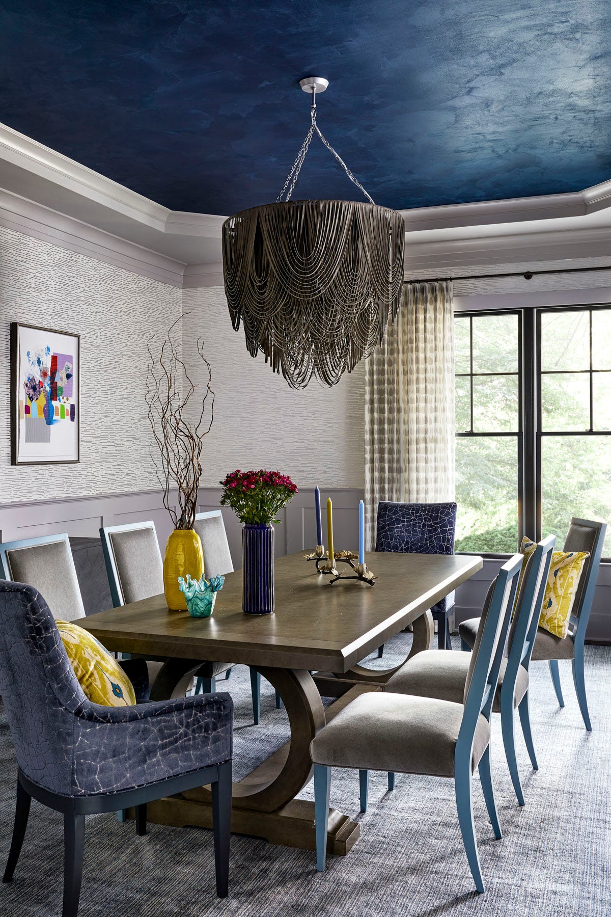 A dining room featuring a dark wood table surrounded by blue and grey chairs under a large metal fringe chandelier and a textured deep blue tray ceiling.