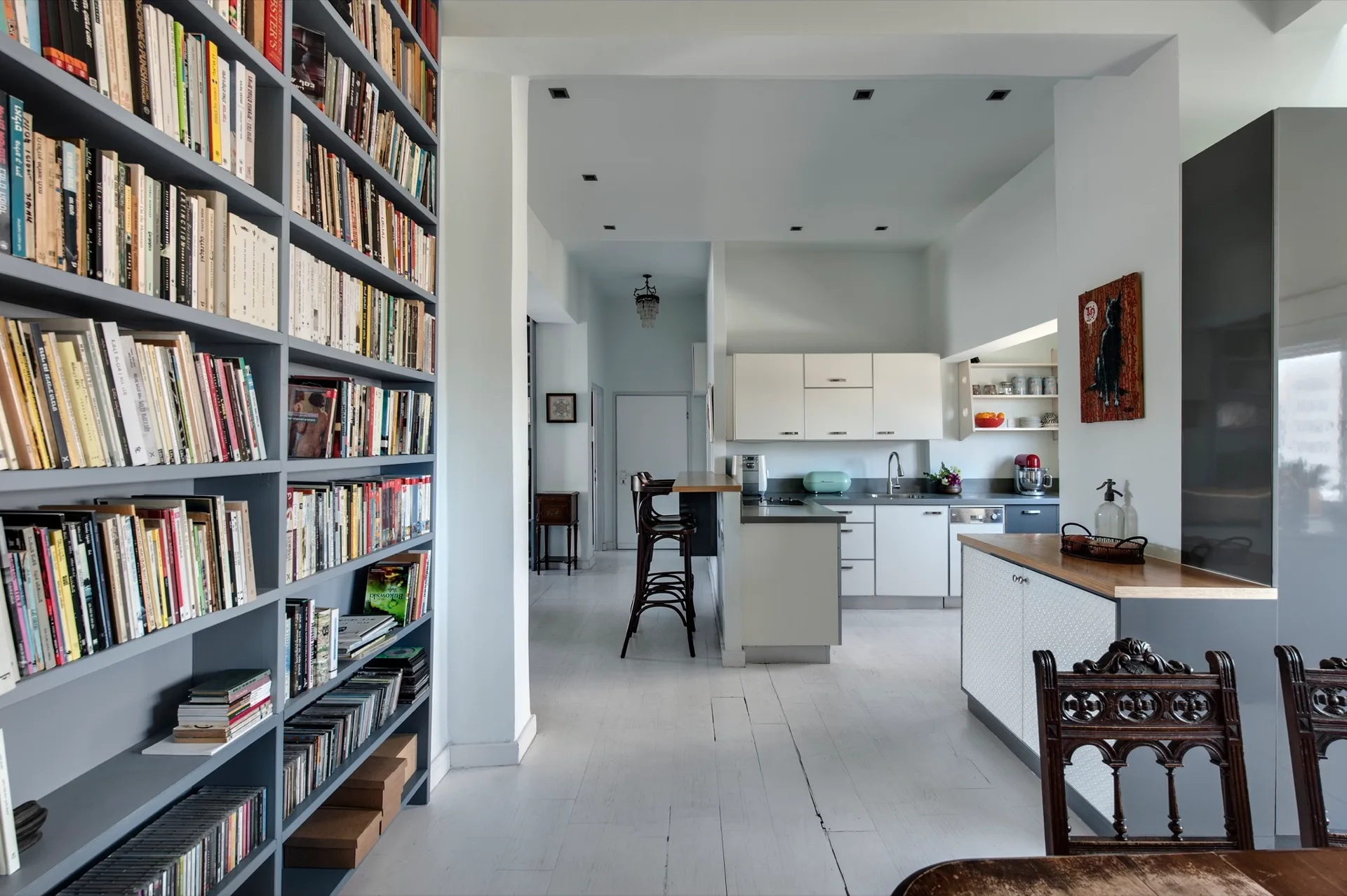 A bright open-plan kitchen features white cabinetry and wooden bar stools alongside a tall grey built-in bookshelf filled with books.