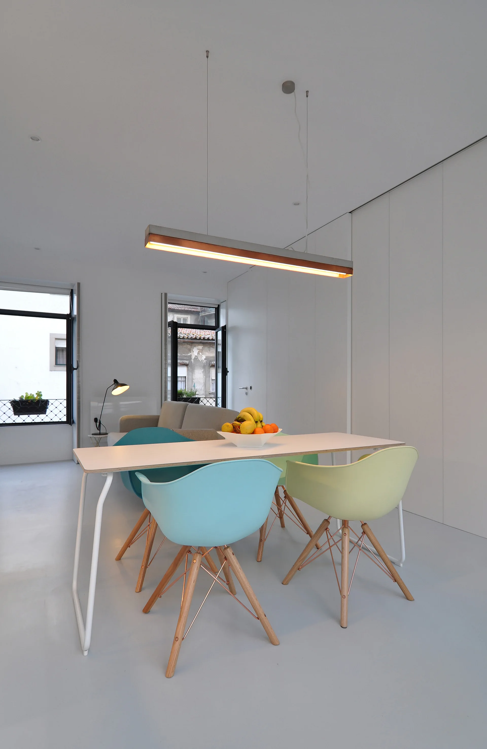A modern dining area features a white table surrounded by teal and pale green molded chairs beneath a glowing linear pendant light.