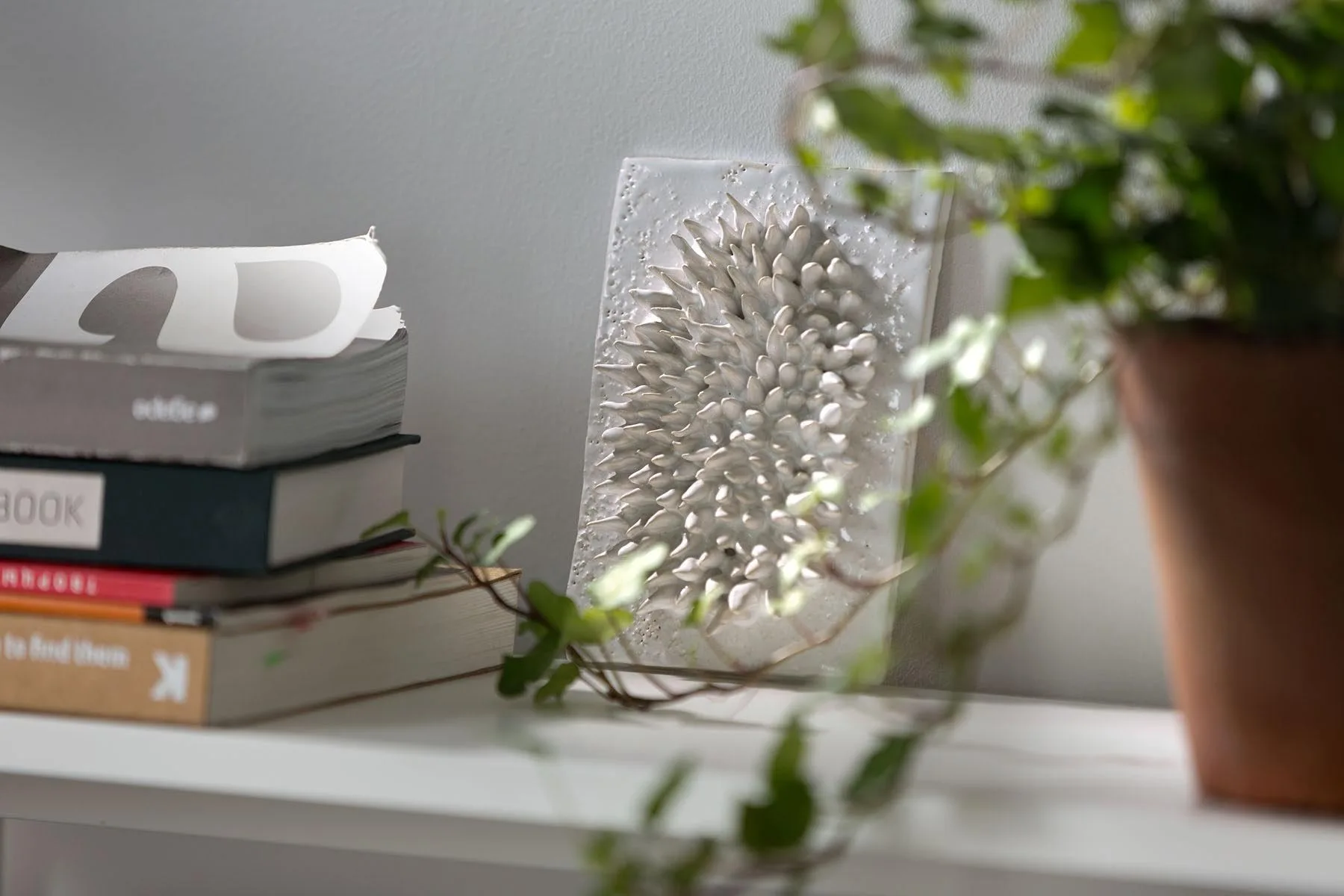 A textured white spiked ceramic art piece rests on a shelf next to a stack of books, with a green potted plant in the foreground.