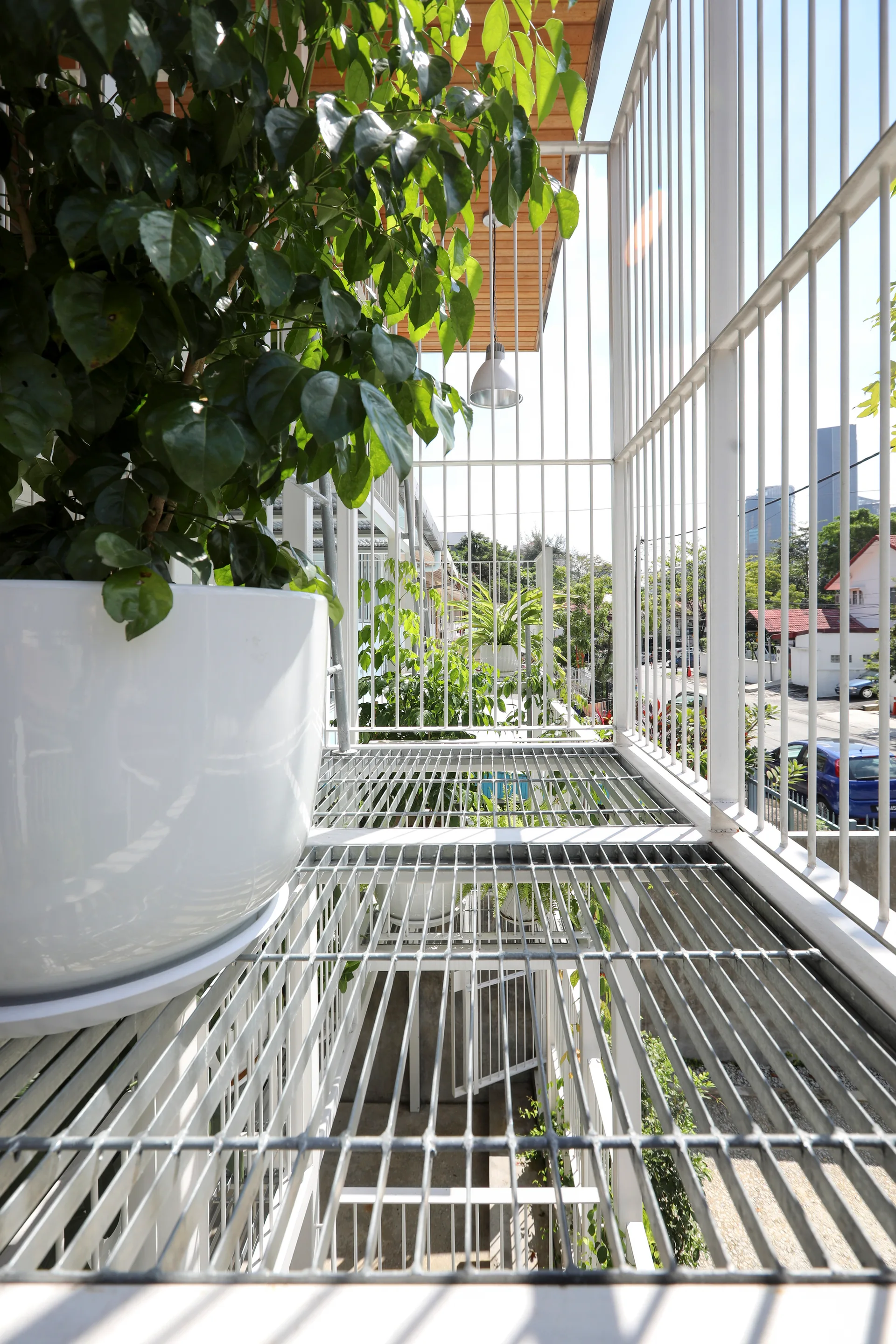 An outdoor industrial balcony features a silver metal grated floor, vertical white steel railings, and a large green plant in a white pot.