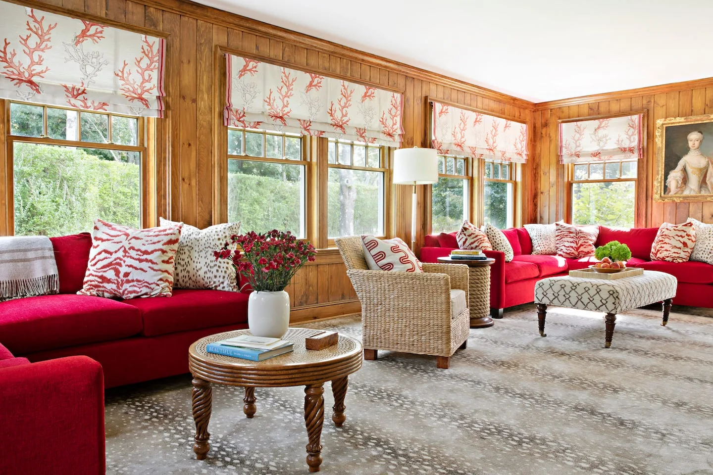 A living room features vertical wood paneling, two bright red sofas, coral-patterned Roman shades, and a woven wicker armchair.