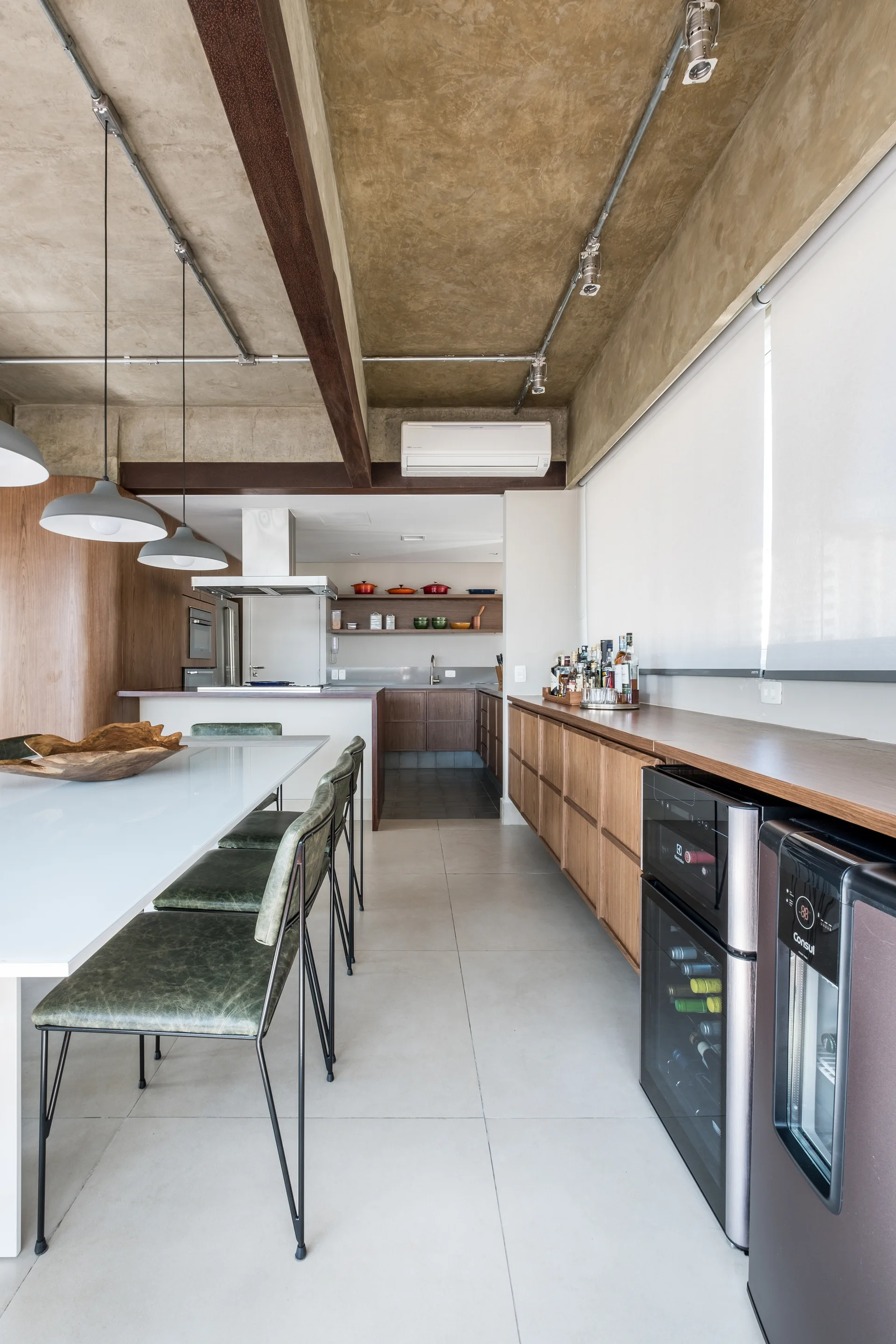 An open-plan kitchen and dining area features an exposed raw concrete ceiling, a white dining table with green chairs, and long wood cabinetry.
