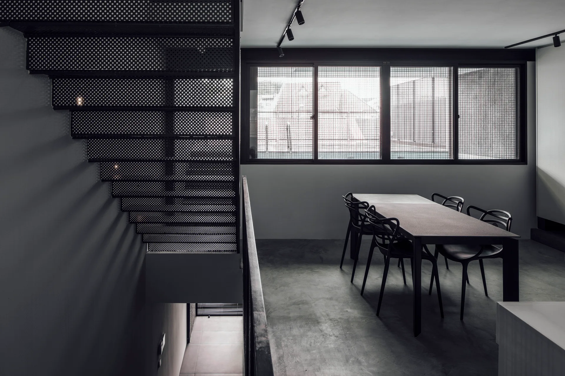 A minimalist dining room features a dark dining table and chairs beneath a perforated metal staircase, with grid windows in the background.
