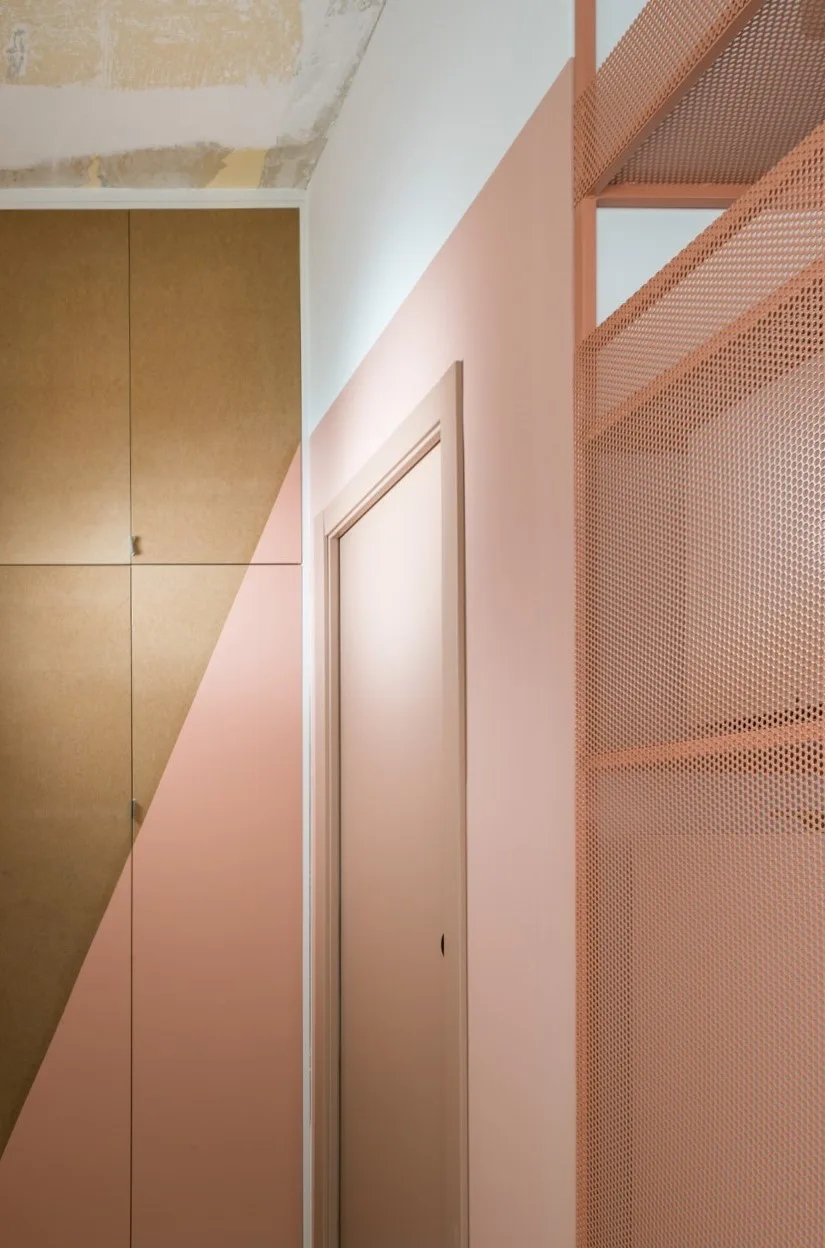 A pink wall with an integrated door sits next to wood-toned tall cabinets painted with a diagonal pink color block and a pink metal mesh shelving unit.