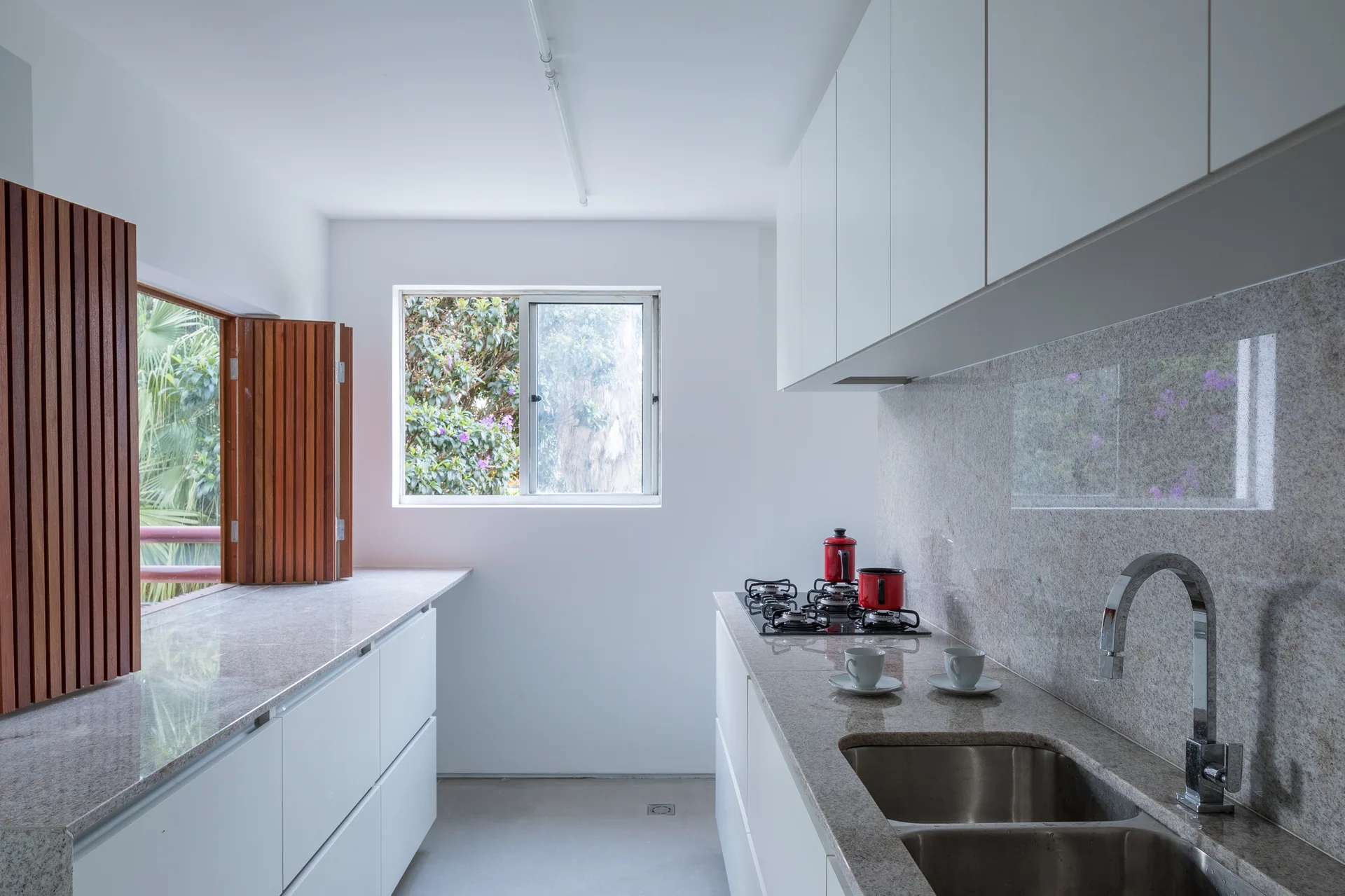 A minimalist galley kitchen features white handleless cabinets, a speckled stone countertop, a steel sink, and louvered wood shutters over a window.