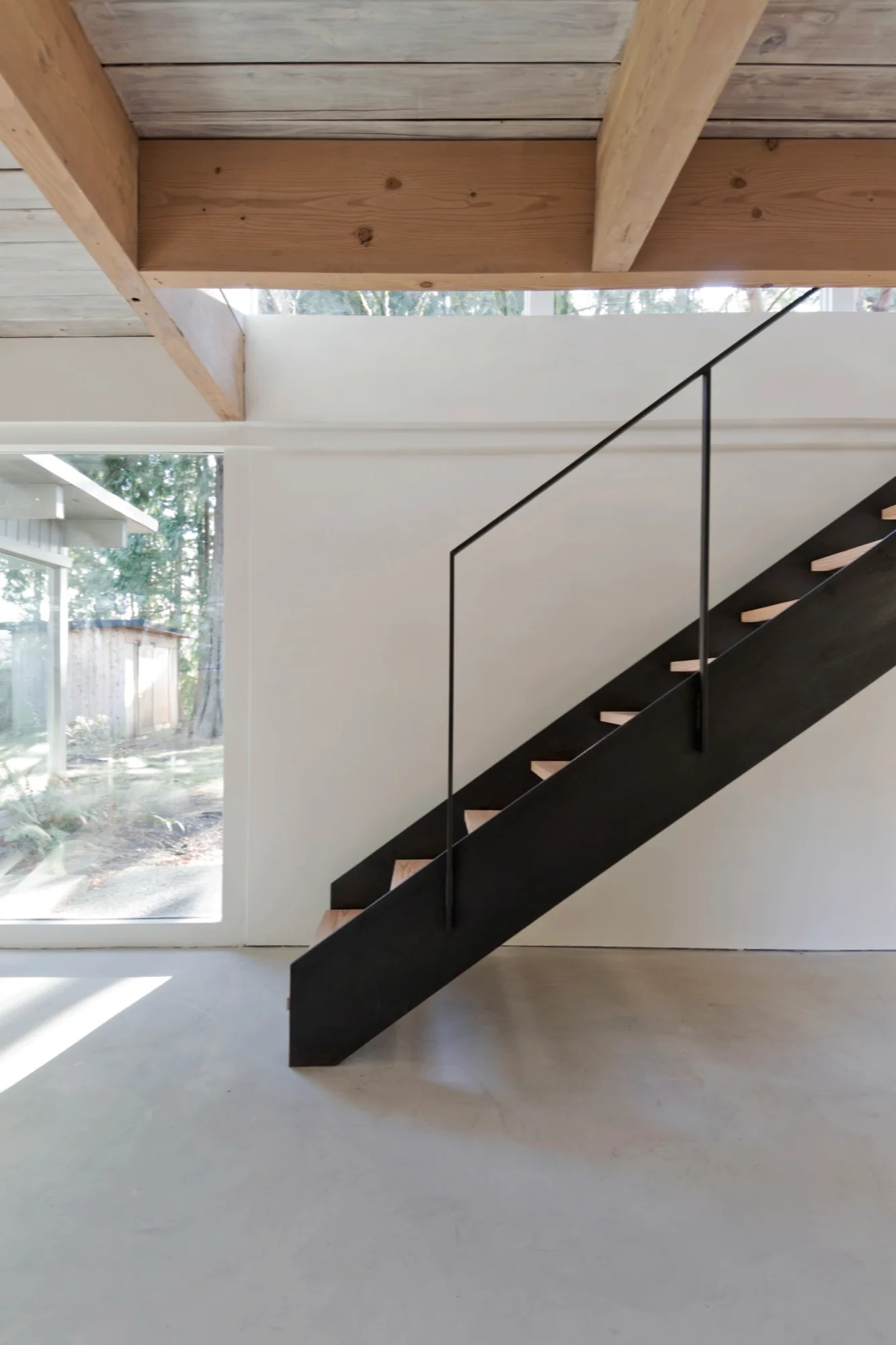 A modern staircase with black steel stringers and wooden treads stands in a room with polished concrete floors and exposed timber ceiling beams.