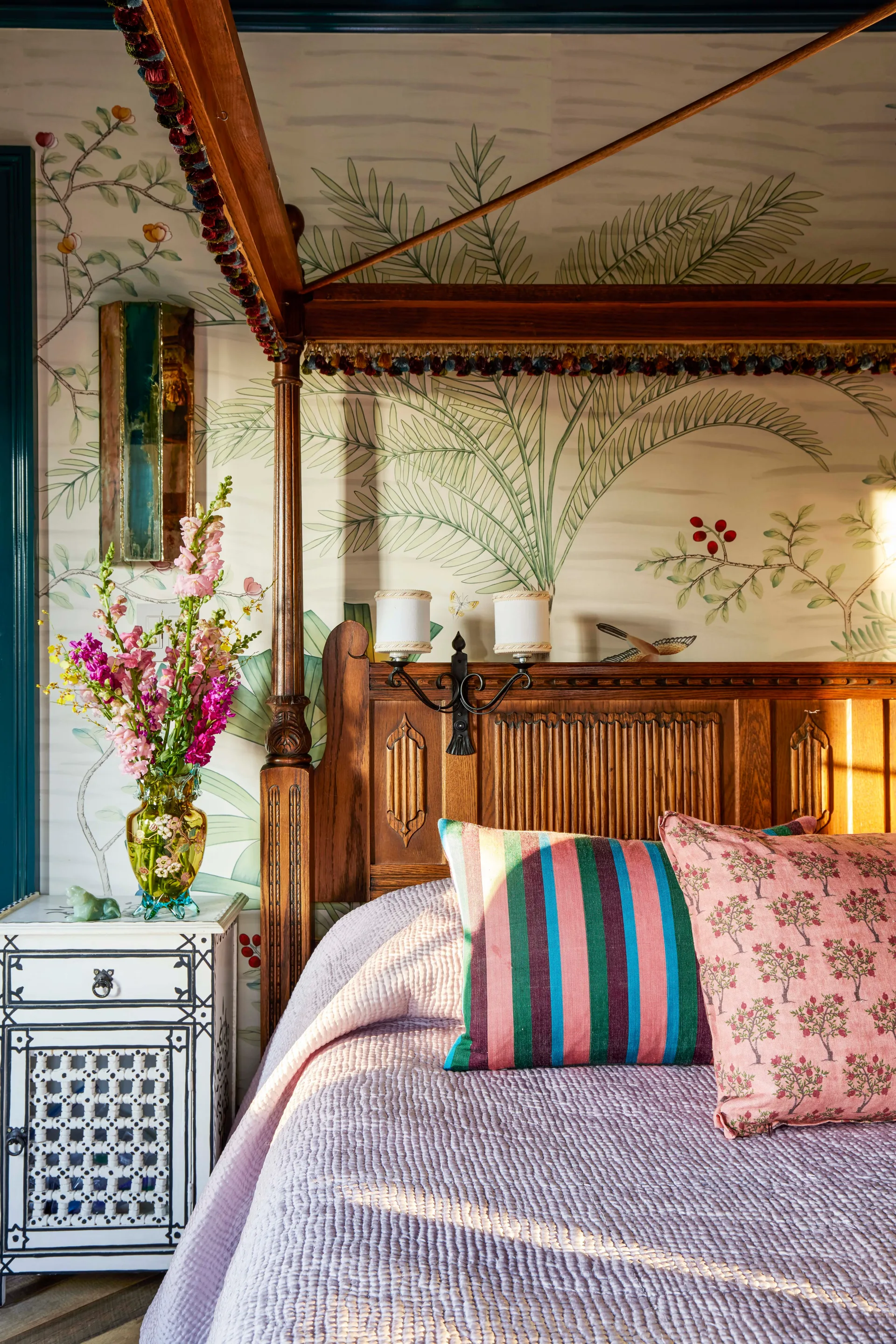 A bedroom corner featuring a carved wooden four-poster bed with colorful bedding against a botanical wallpaper pattern and next to an inlaid nightstand.