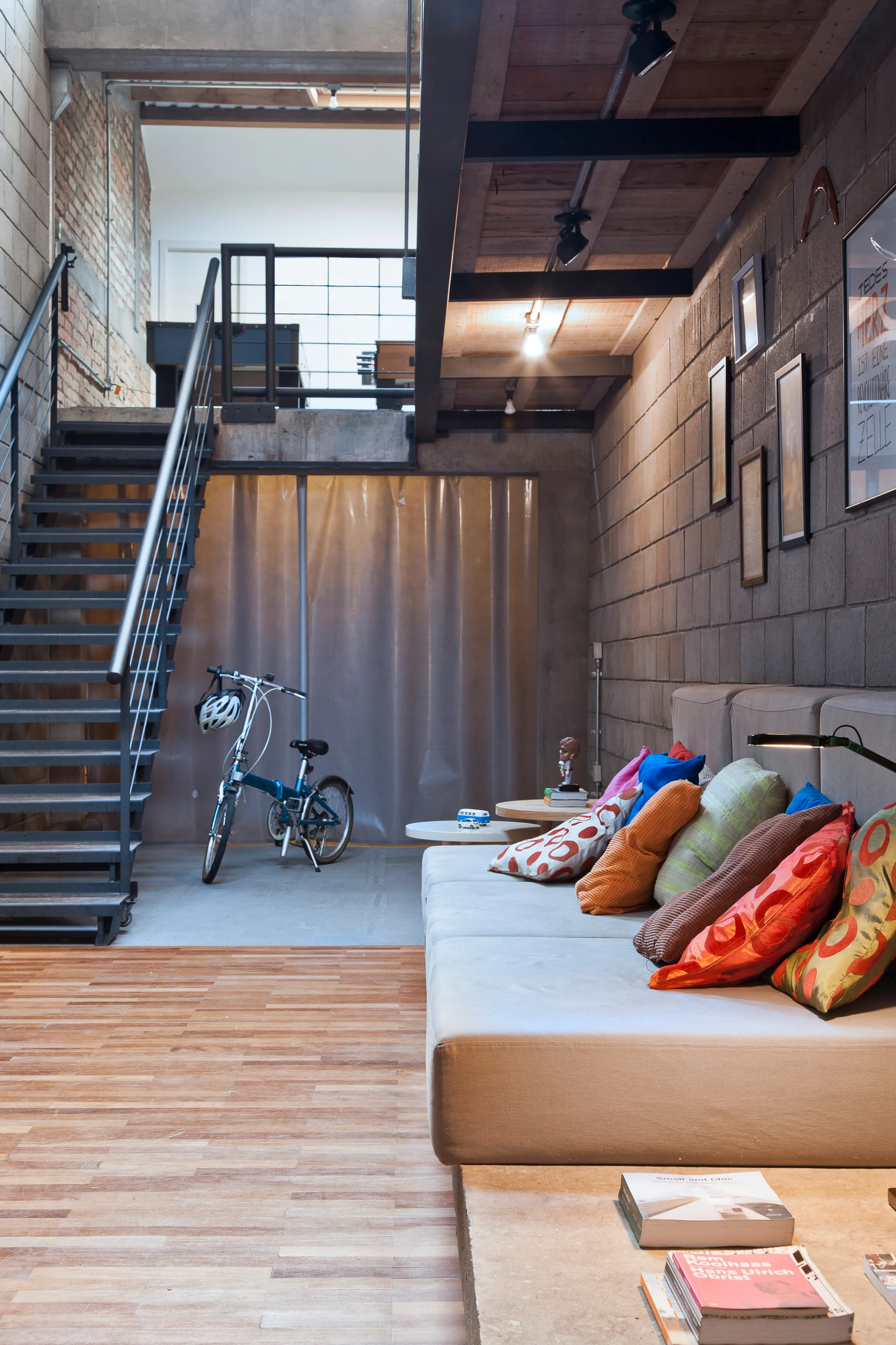 An industrial loft living room with concrete block walls, a low gray sofa on a concrete platform, colorful pillows, and a metal staircase.