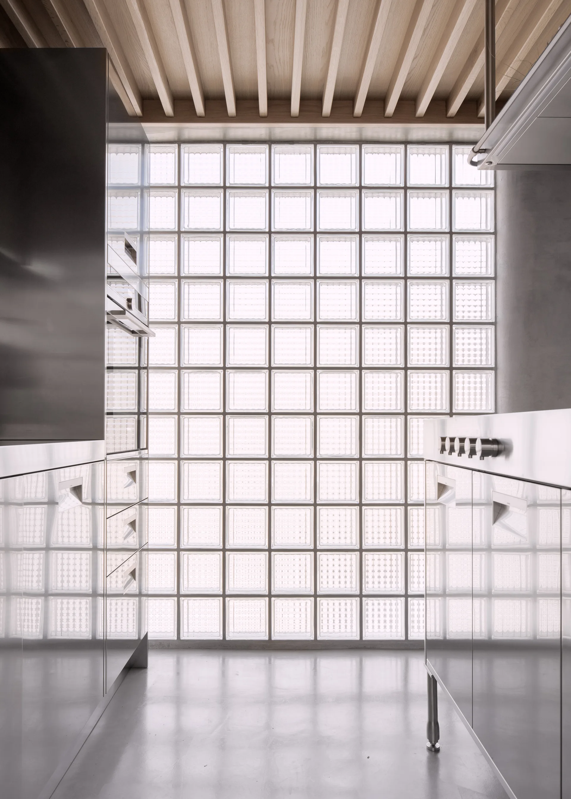 A modern kitchen featuring reflective stainless steel cabinetry on both sides, a concrete floor, a timber slat ceiling, and a large wall of glass blocks.