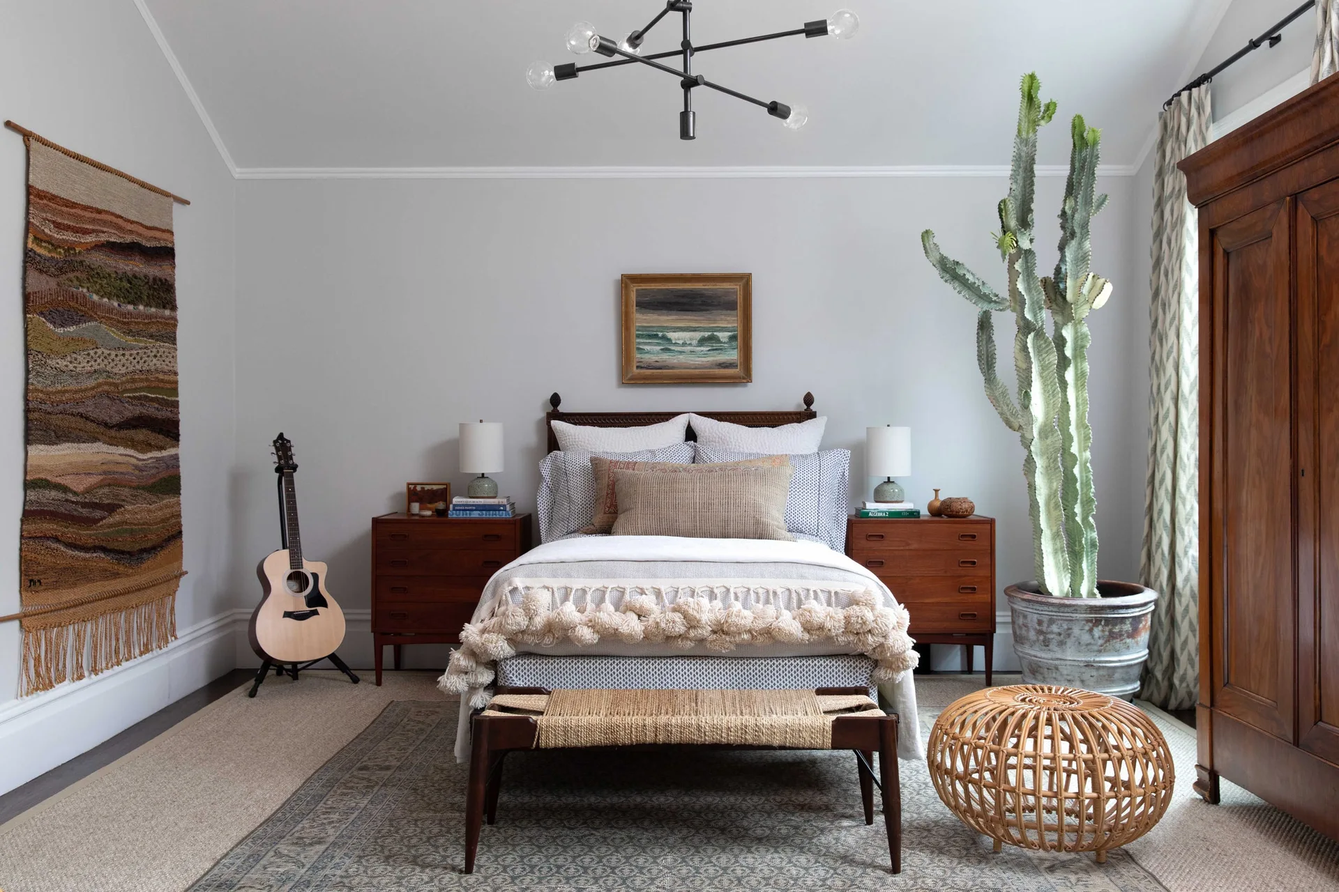 A bedroom features a bed with neutral bedding, mid-century wood nightstands, a large potted cactus, and a textured wall hanging.