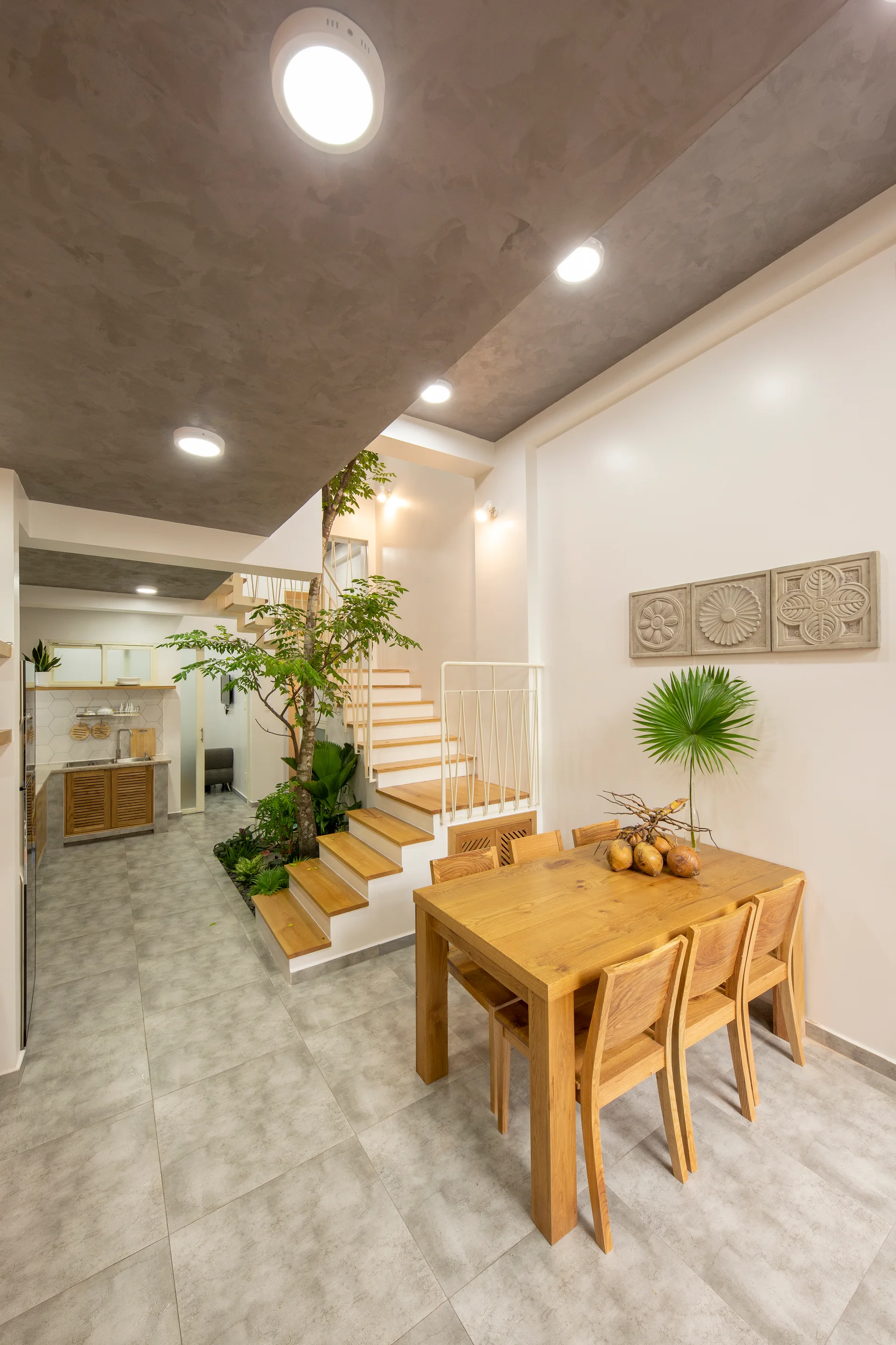 A light wood dining table and chairs stand near a white staircase with an integrated indoor garden bed and grey tiled floor.