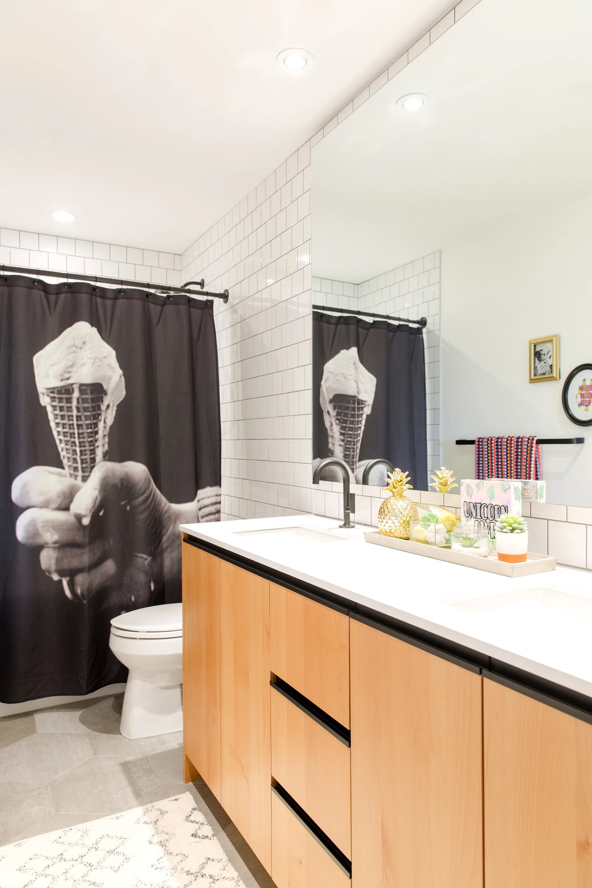 A modern bathroom features a light wood vanity with a white countertop, matte black faucets, white square tiled walls, and a black-and-white shower curtain.