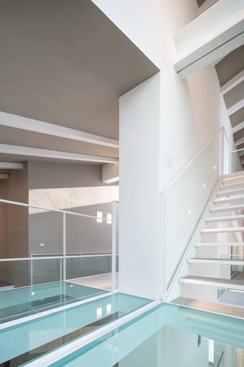 A minimalist white metal staircase ascends next to a transparent structural glass floor bridge under a taupe ceiling with exposed white beams.