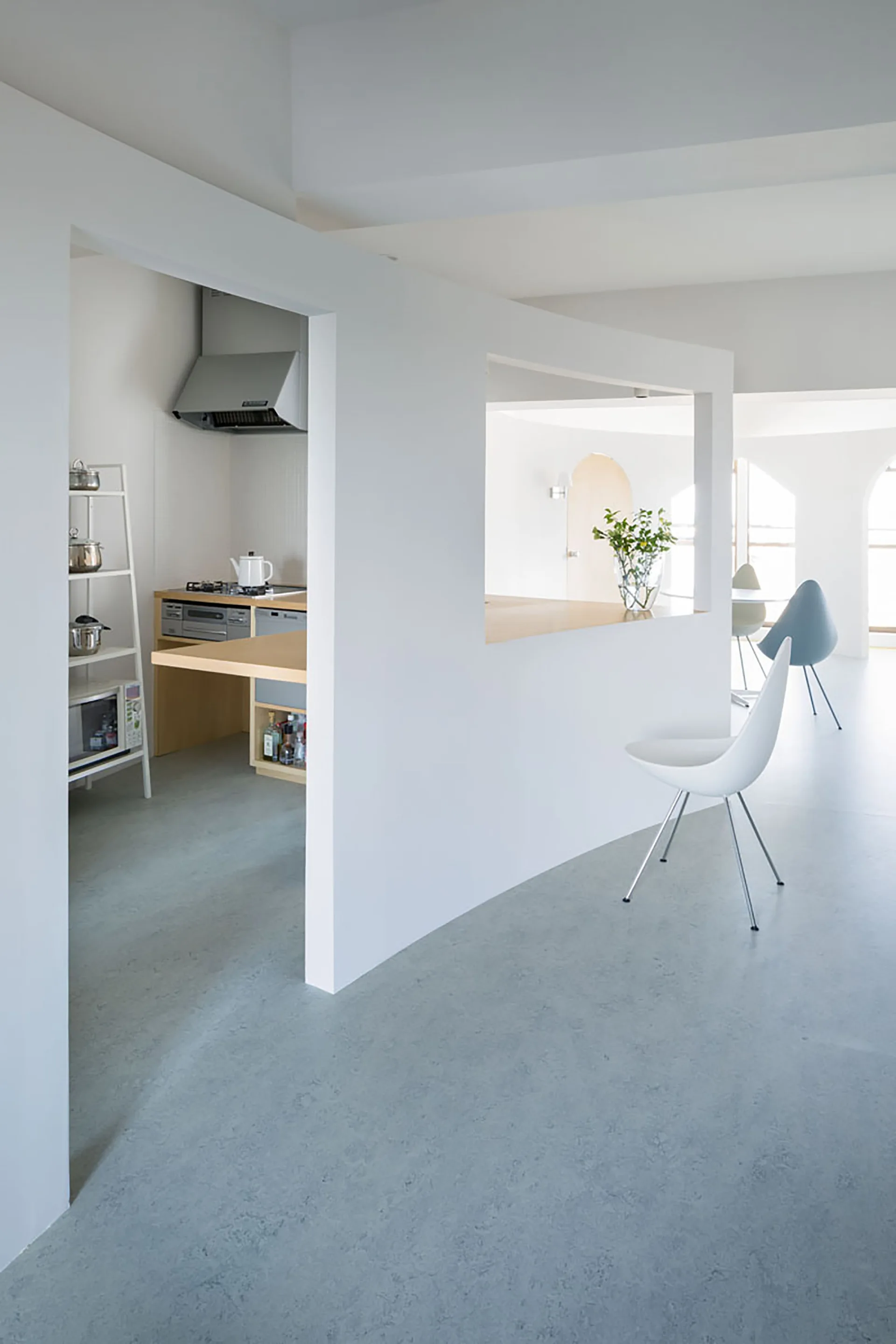 A white partition wall with a rectangular cutout separates a minimalist kitchen from an open dining area featuring molded chairs and a smooth gray floor.