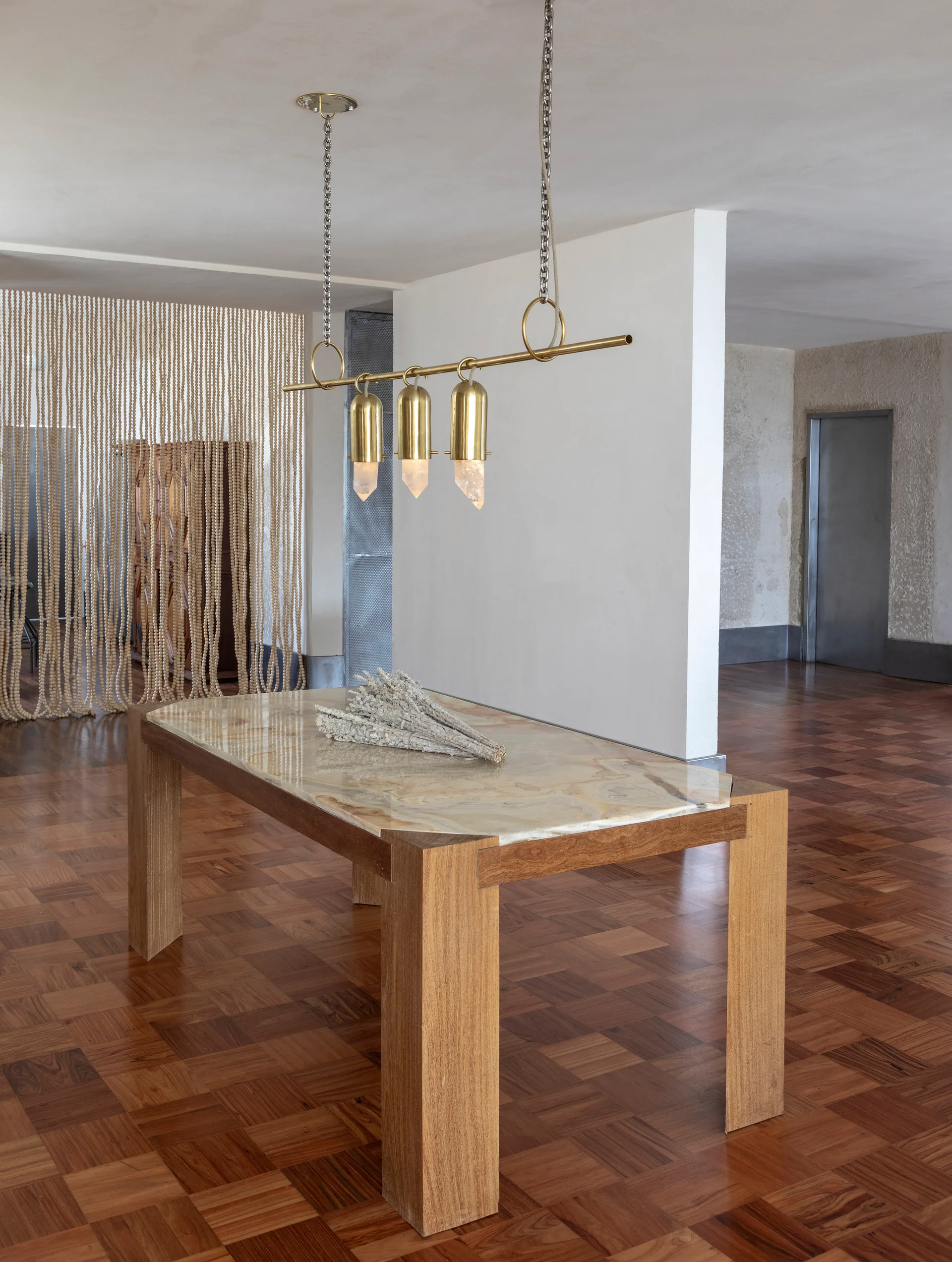 A wooden dining table with a polished stone top sits beneath a brass and crystal pendant light in a room with parquet flooring and a beaded screen.