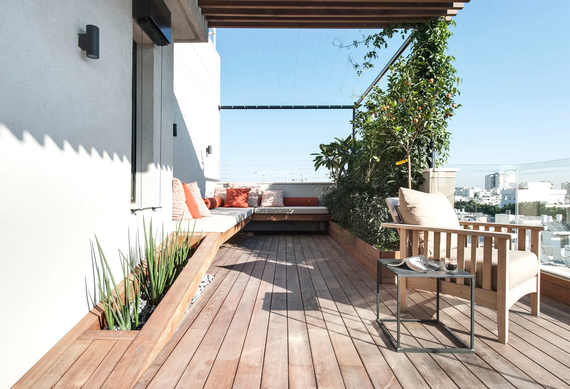 A modern rooftop terrace features warm wood decking, an L-shaped built-in bench with orange pillows, and a recessed floor planter with snake plants.