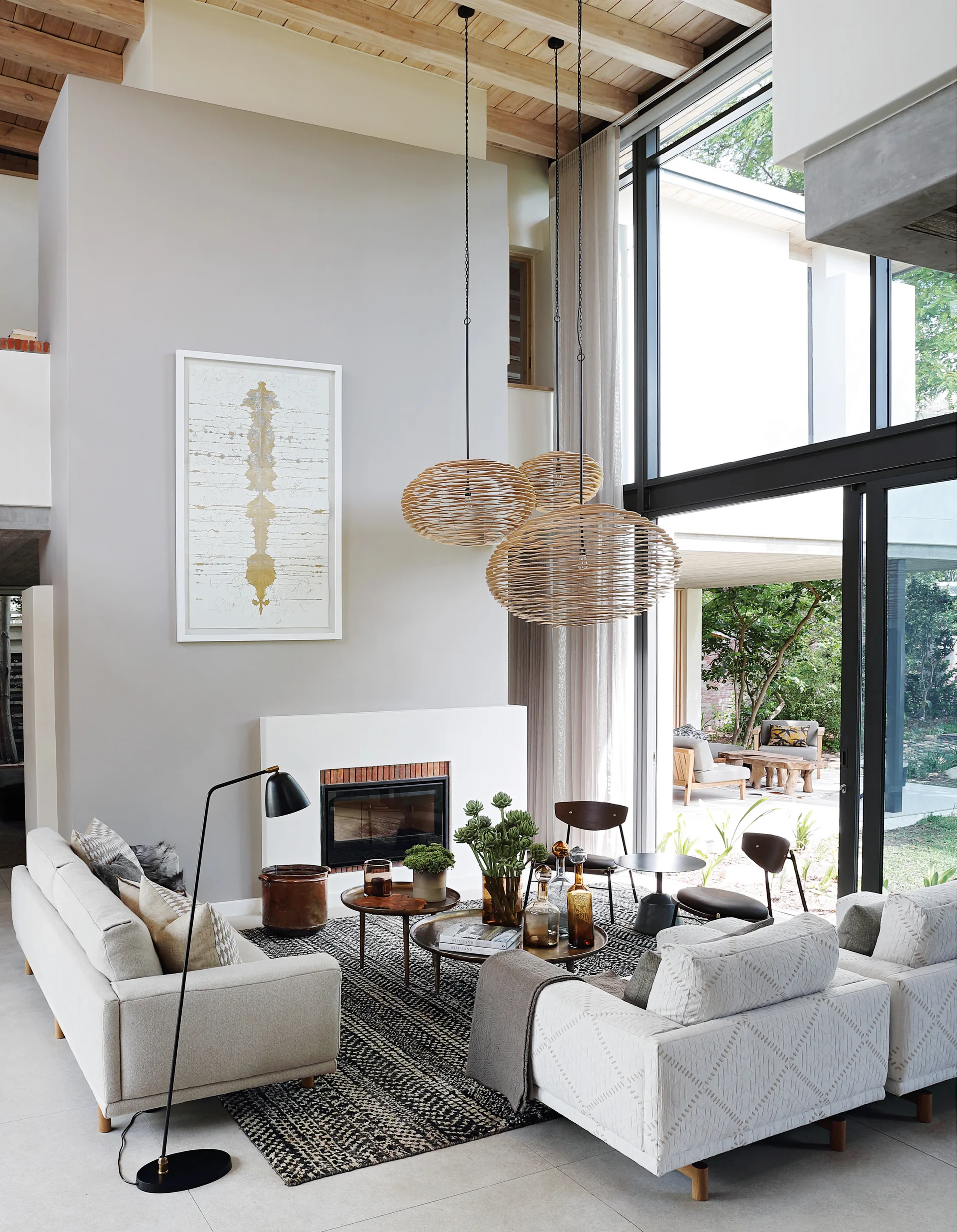 A double-height living room features light grey sofas, a patterned rug, nested wooden coffee tables, and three woven globe pendant lights near a fireplace.