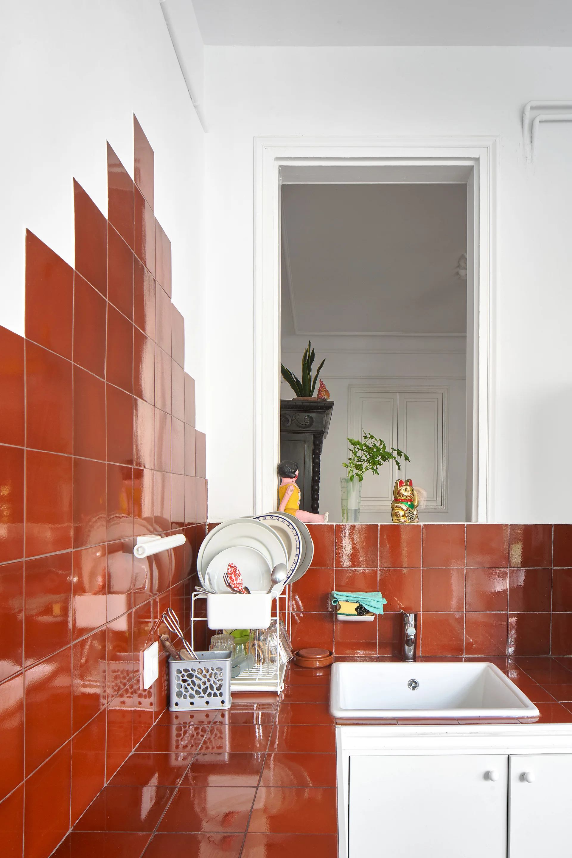 A kitchen features a glossy red-brown square tiled backsplash and countertop, a white ceramic sink, and a pass-through opening to another room.