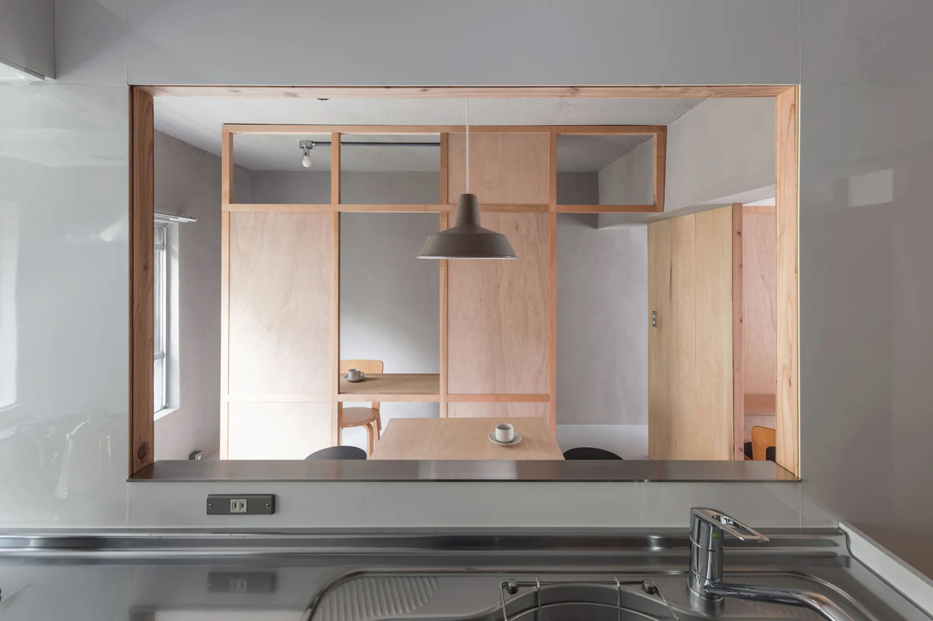 A view from a stainless steel kitchen sink looks through a wood-framed opening to a dining area with a light wood partition and a grey pendant light.
