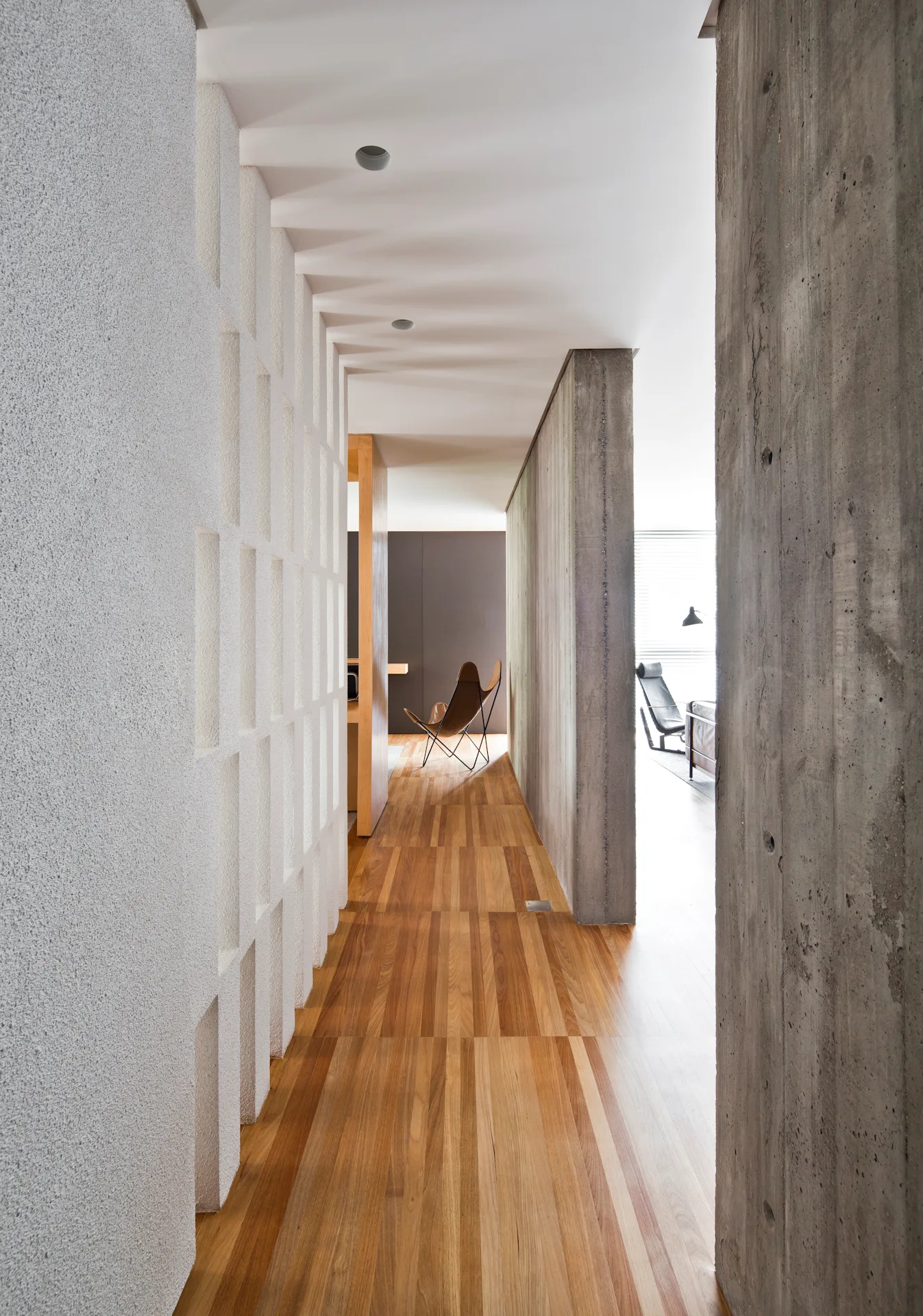A modern hallway features a white plaster grid wall, raw concrete columns, and warm wood flooring leading to a sunlit living space.