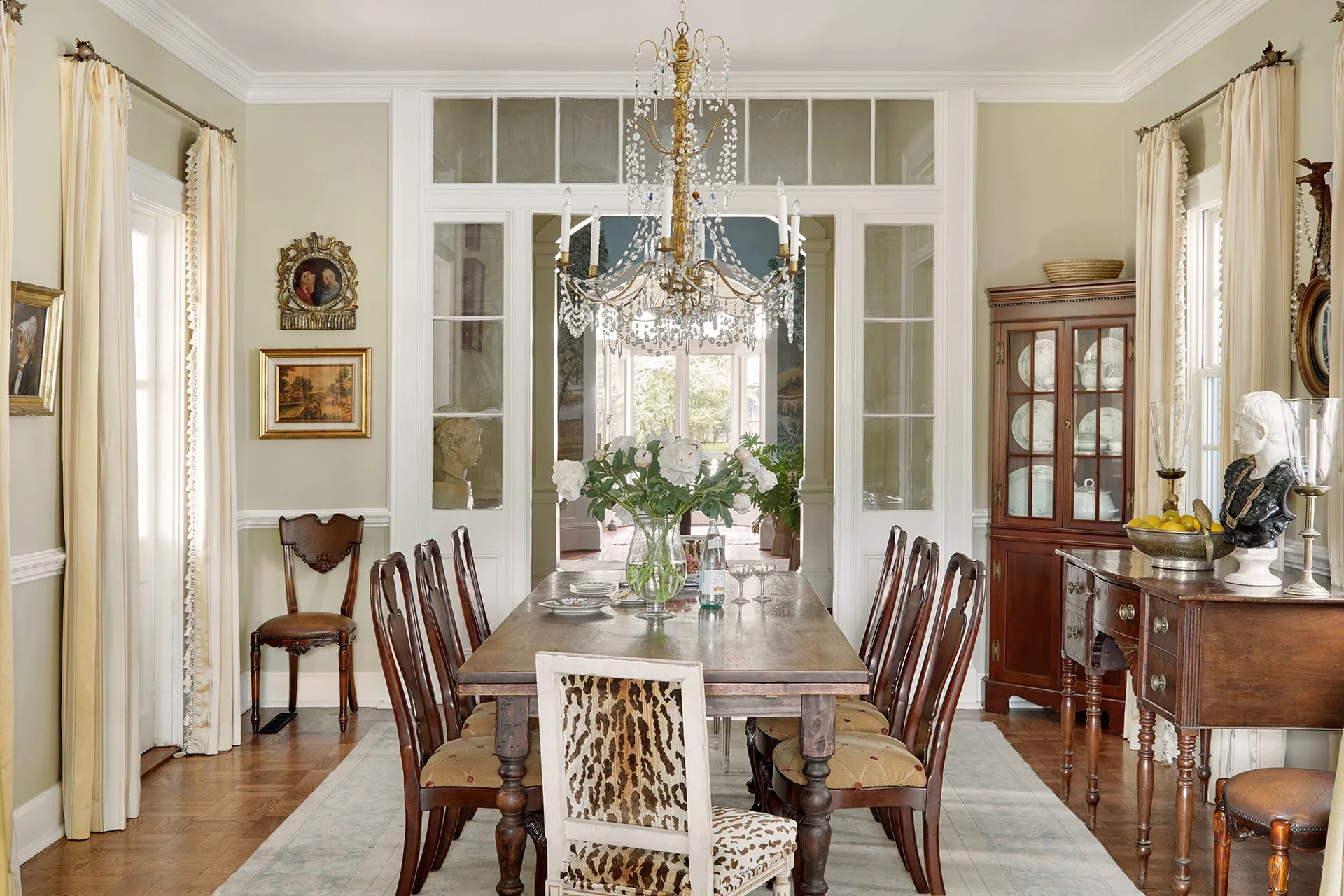 A formal dining room features a dark wood dining table, sage green walls with white trim, a crystal chandelier, and a corner china cabinet.