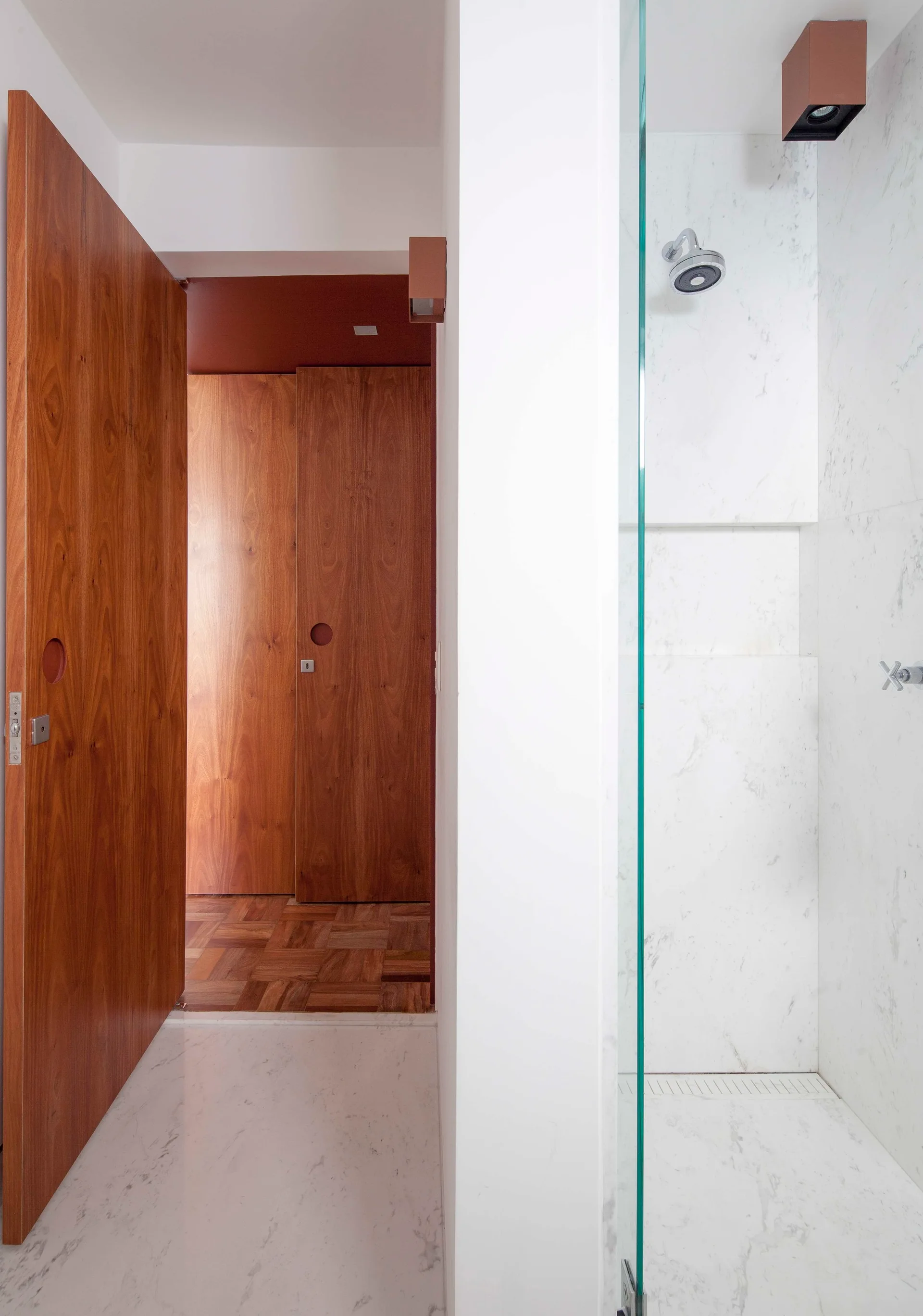 A glass-enclosed white marble shower stands next to a hallway with warm wood doors and parquet flooring.