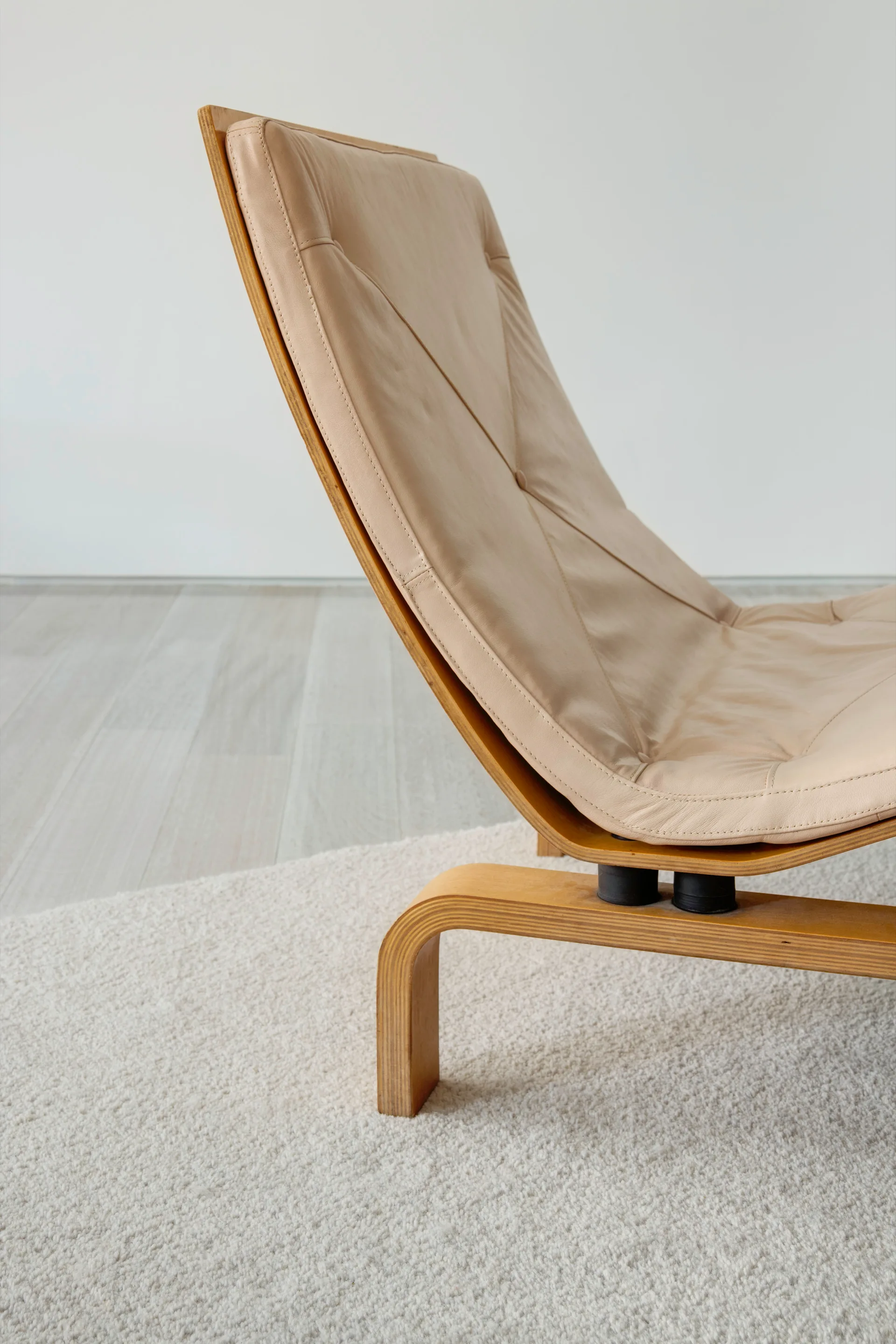 A beige leather lounge chair with a molded plywood frame sits on a textured off-white rug next to a white wall.