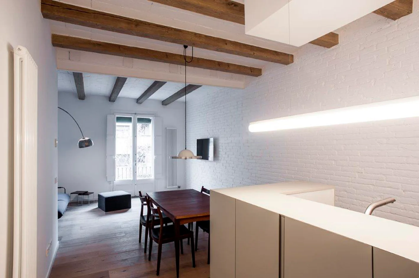 A modern open-plan dining space features a wooden table, exposed timber ceiling beams, and a white painted brick wall.