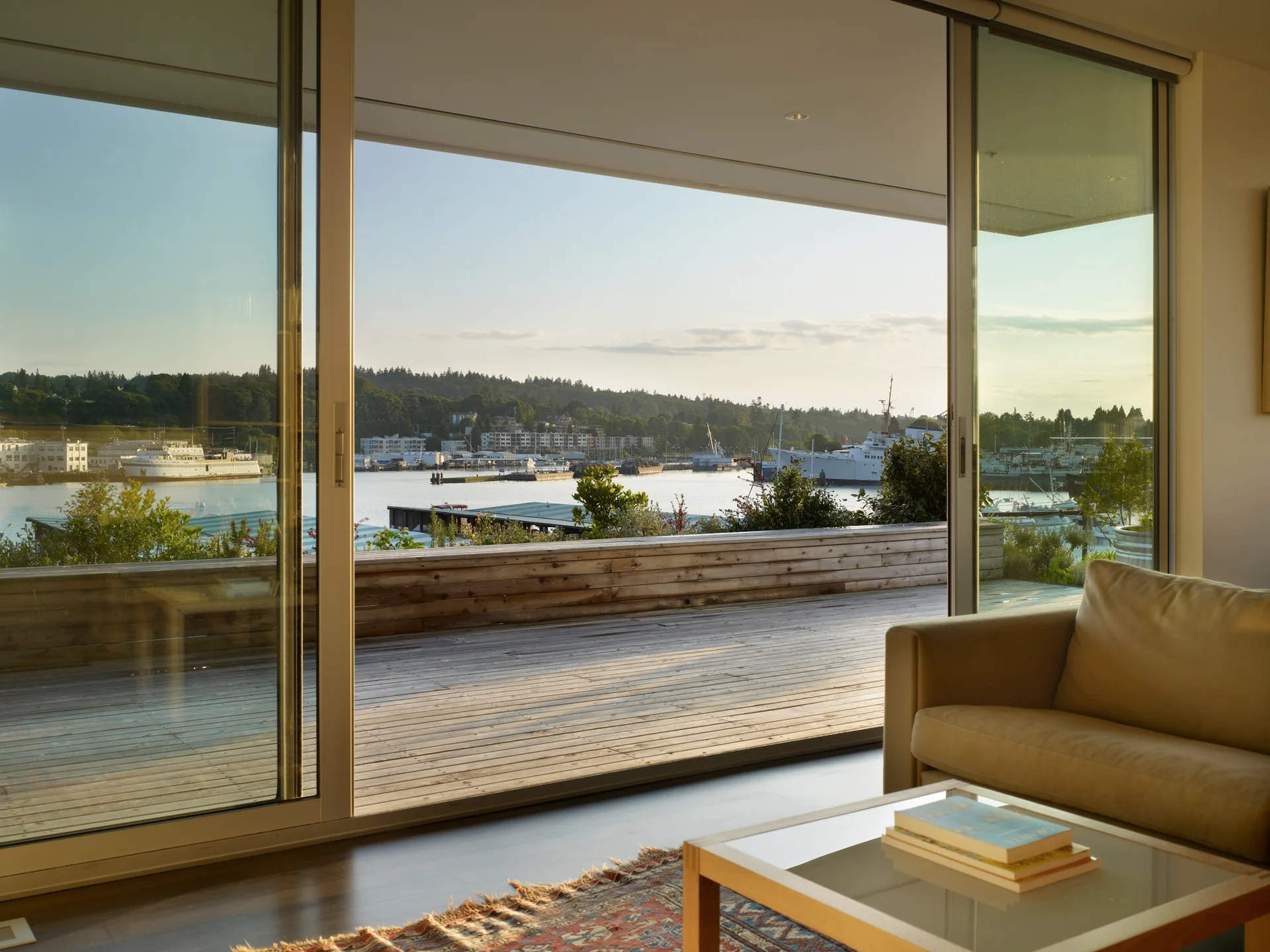 A living room with a beige sofa and patterned rug features wide sliding glass doors that open to a wood deck overlooking a harbor.